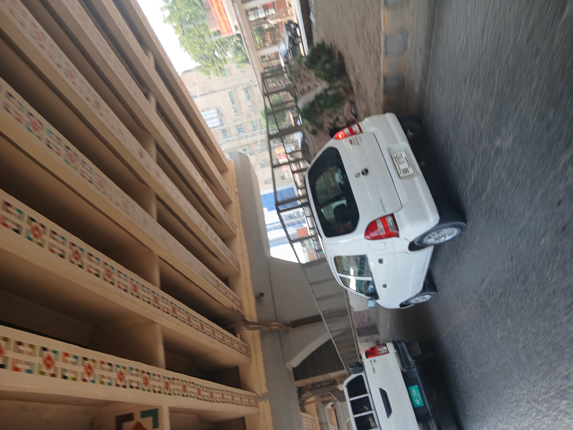 The image depicts a street scene in Peshawar City Tehsil, Pakistan, beneath an elevated structure, possibly a bridge or overpass. The undercarriage of the structure is visible, featuring decorative bands with geometric patterns in red, blue, yellow, and white. Two white vehicles are the primary subjects. In the foreground, a white pickup truck is positioned on the left, its rear end facing the camera. To its right, a white hatchback is seen from its rear quarter, angled towards the right. Both vehicles appear to be in motion or parked on a paved road. In the background, the environment includes buildings, trees, and other vehicles, suggesting an urban or semi-urban setting. The lighting suggests it is daytime. The focus is on the vehicles and the overhead structure.