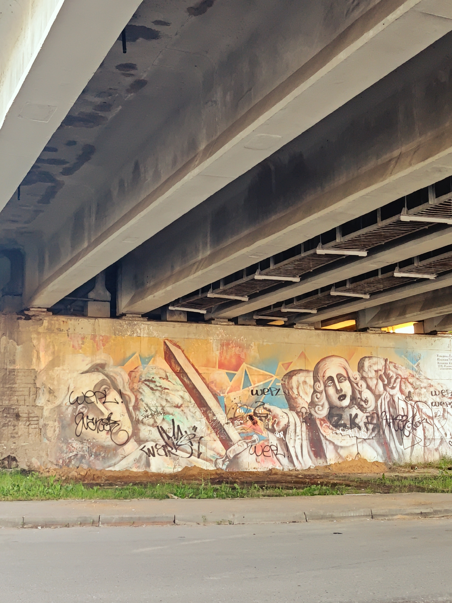 This image depicts a section of a concrete overpass in Kimry, Russia, with a large graffiti mural painted on the retaining wall beneath it. The overpass is constructed with multiple parallel concrete beams, some of which show signs of water staining and discoloration. The retaining wall is weathered and appears to be made of brick and concrete.
The graffiti mural features several figures, predominantly faces with exaggerated features. The central figure is a woman with wide eyes and an open mouth, surrounded by other figures that appear to be partially rendered or in the background. The style is somewhat abstract and unsettling. The mural is also covered with various tags and signatures in black spray paint, including "werz" and other illegible markings.
In front of the wall, there is a strip of green grass and weeds, followed by a paved surface, likely a road or sidewalk, with a curb. The lighting suggests it is daytime, with warm light filtering from the right side of the frame, possibly indicating the setting sun. The overall scene is urban and somewhat neglected, characterized by the juxtaposition of the massive concrete structure and the vibrant, albeit graffiti-laden, artwork on the wall.