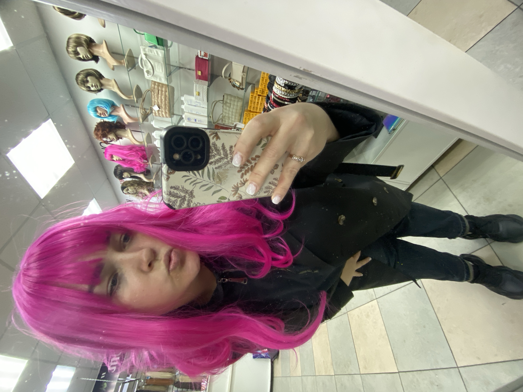 A person with bright pink, shoulder-length hair and bangs, possibly a wig, takes a mirror selfie in what appears to be a retail store. The person is wearing a dark, possibly black, jacket or coat, dark jeans, and black boots. They hold a smartphone with a distinctive white case featuring a brown and green floral or leafy pattern in their right hand. This hand also displays fingernails with white tips (French manicure) and a ring on one finger. The person's face is visible, showing a slight pout.
The background, seen as a reflection in the mirror, reveals several display shelves. The top shelves feature multiple mannequin heads wearing various wigs in different colors and styles, including blonde, blue, brown, and pink. Below these, more shelves hold an assortment of merchandise, including handbags in colors like white, beige, green, and red, alongside other packaged items that appear to be cosmetics or beauty products. Stacks of bracelets or bangles are also visible on a display. The floor of the store is tiled with alternating light and darker square patterns. The indoor lighting is bright, with some reflections visible on the mirror surface. No legible text is discernible on any of the products or signage in the background. The location context provided is Sochi, Russia.