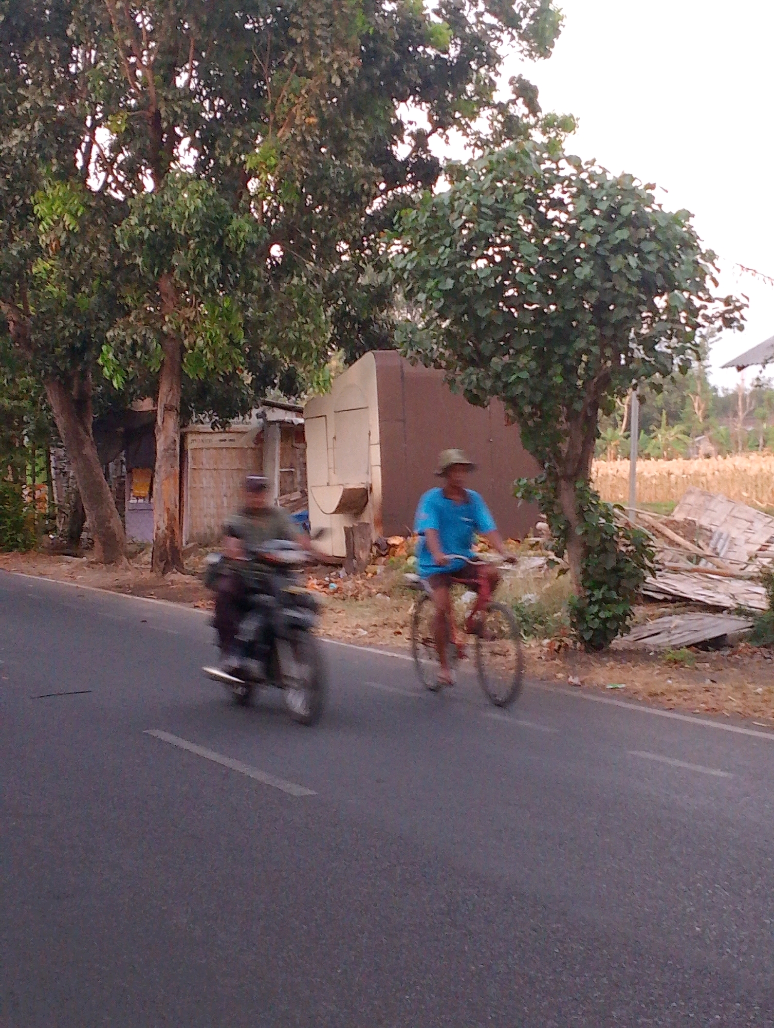 The image depicts a rural road scene in Probolinggo, Indonesia, likely during late afternoon or early morning given the soft light. A person rides a motorcycle on the left side of the road, their form blurred due to motion. On the right, another individual is cycling on a bicycle, also appearing somewhat blurred. They are both on an asphalt road with a white painted line marking the center. The background features lush green trees, a modest building with a metallic exterior, and a field of dry crops. Debris, including wooden planks, is visible on the roadside. The sky is overcast.
