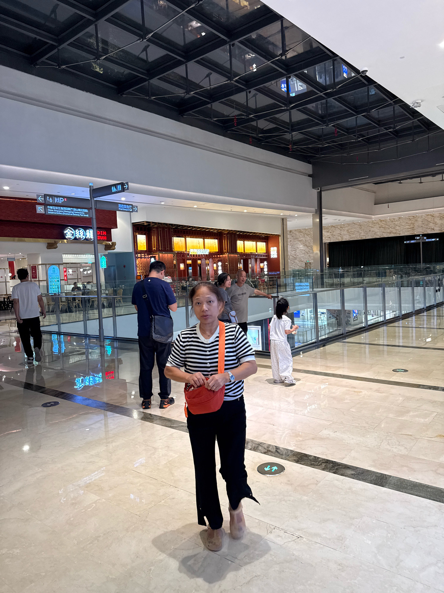 A mid-shot, eye-level full body photograph shows an Asian woman in the foreground, centered within a large, modern indoor shopping mall. The woman is facing the camera, her expression neutral, and she has short, dark hair and is wearing a black and white striped t-shirt, black pants, and beige slippers. She is carrying a bright orange crossbody bag. In the background, several other people are visible, including a man walking away from the camera to the left, a couple standing and looking to the right, and a young girl in a white dress walking away to the right. The mall environment features polished marble flooring with dark inlaid lines, a multi-level structure with railings and glass partitions, and a dark, grid-like ceiling with lights. Signage is visible in the background, including directional signs in Chinese and English, and the name of a restaurant, "DIM SUM". The overall impression is a brightly lit, clean, and spacious commercial area in Yuzhong District, China.