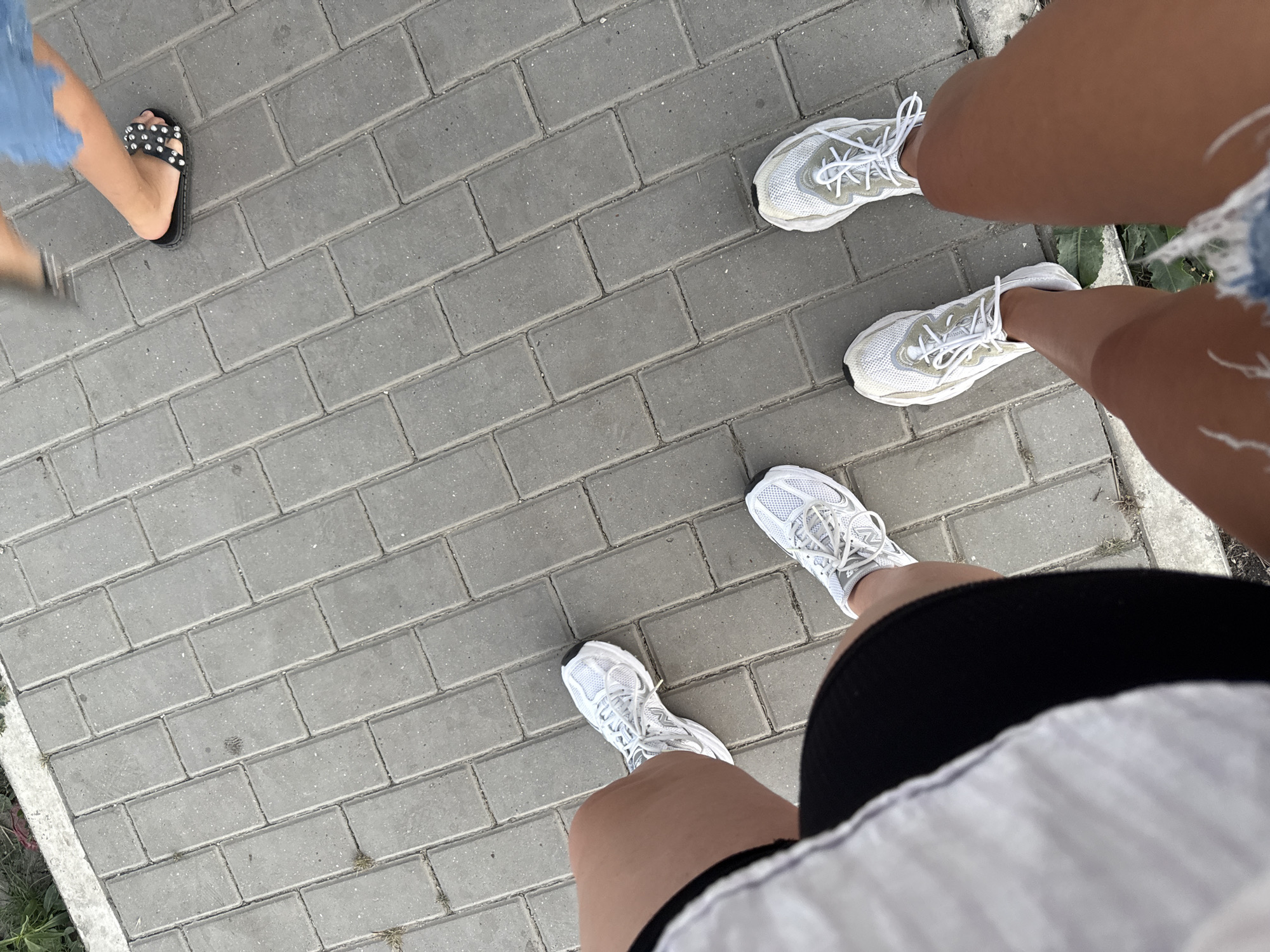 This is a point-of-view shot of two pairs of legs and feet on a grey brick pathway. The feet on the left are wearing white athletic shoes. The legs on the left are wearing black shorts. The feet on the right are wearing white athletic shoes. The legs on the right are wearing ripped denim shorts. In the upper right corner, a third person's legs and feet are visible, wearing black slide sandals. The pathway is made of rectangular grey bricks laid in a running bond pattern. Some sparse green vegetation is visible at the edges of the pathway. The scene is outdoors.