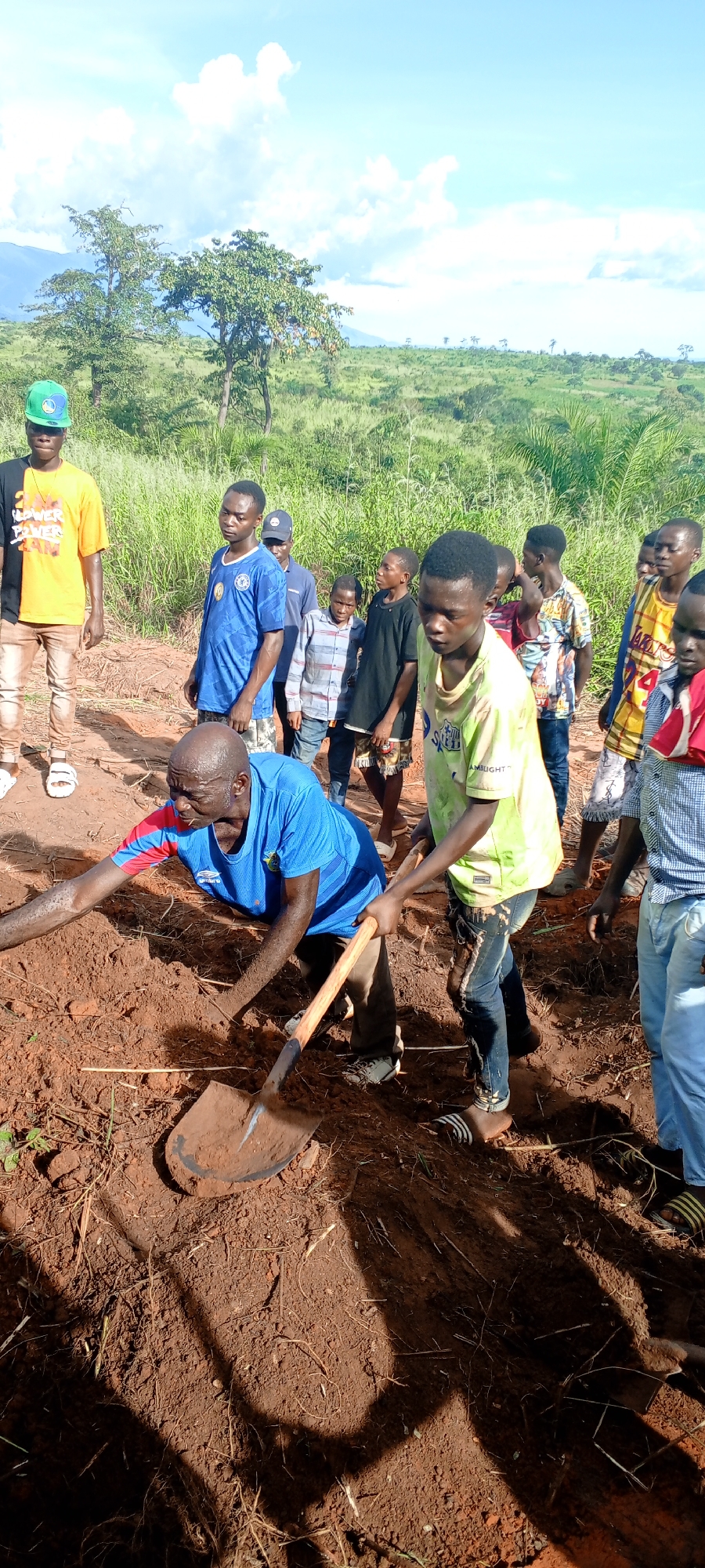 In Ngalula, Democratic Republic of the Congo, a group of individuals is gathered outdoors in a grassy, somewhat rural location under a partially cloudy sky. A man in a blue shirt is digging into the reddish-brown soil with a shovel. A younger person in a light green shirt assists him. Other people stand nearby, observing the digging. The landscape shows trees and tall grass. The setting has a rural feel, with open land.