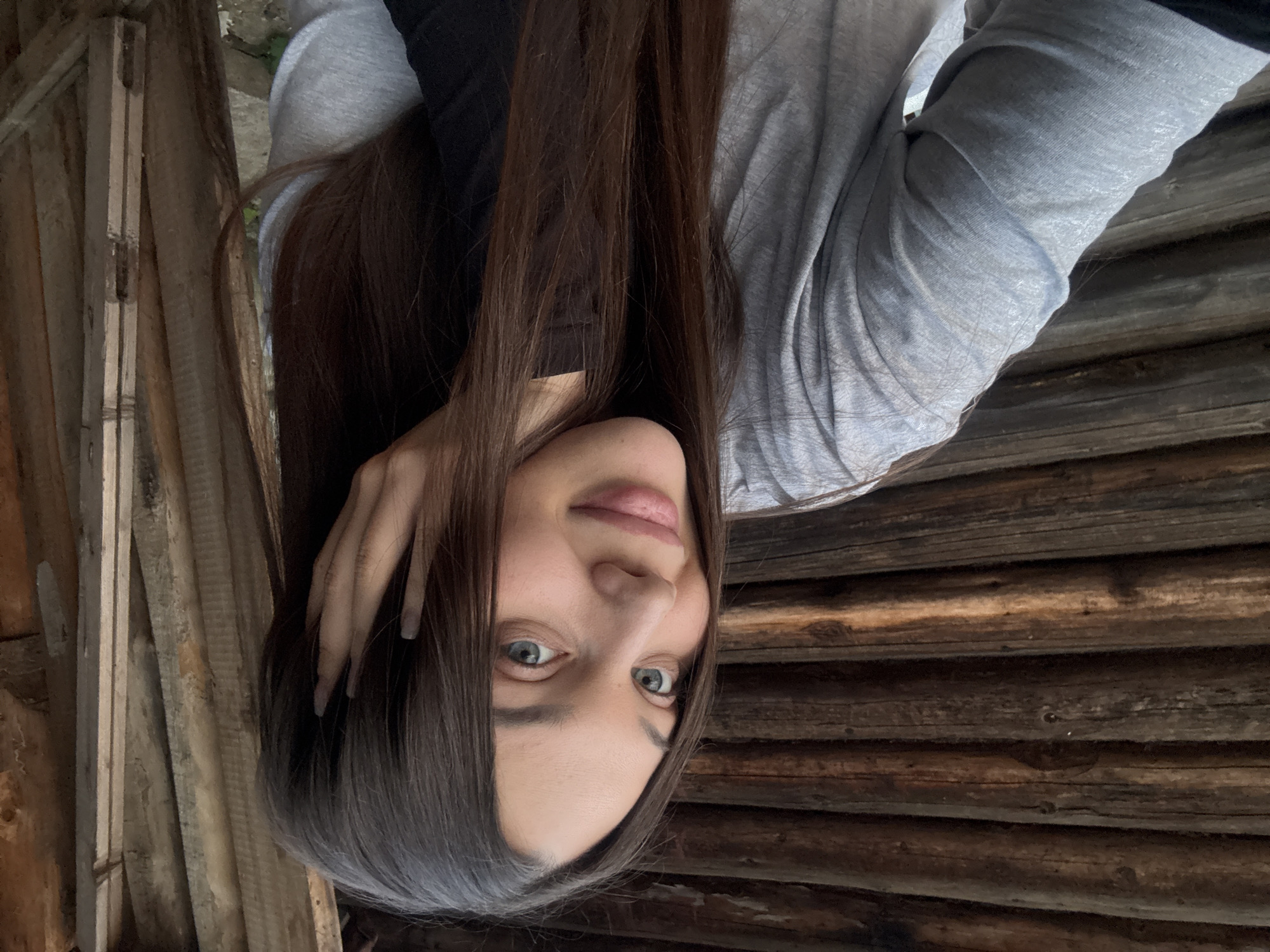 A woman with long brown hair and blue eyes poses against a wooden wall. She is wearing a gray long-sleeved shirt over a black top. Her hand is touching her hair. The background appears to be a wooden structure, possibly an outbuilding or shed. The location is Chaikovsky, Russia.