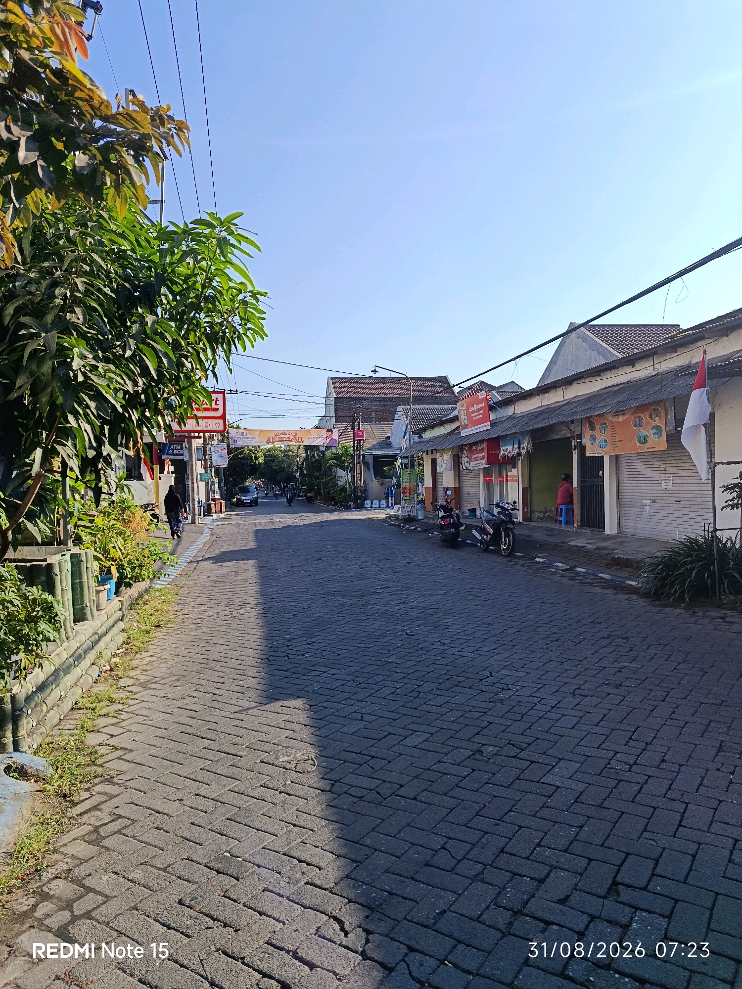 The image depicts a street scene in Keboansikep, Indonesia, under a clear blue sky. The street is paved with dark gray interlocking bricks laid in a herringbone pattern. On the left side of the street, a lush green tree with dark leaves overhangs the sidewalk, which is bordered by a low fence made of bamboo poles. Beyond the fence, there are some green plants and a blue concrete object.
The right side of the street features a row of single-story buildings with tiled roofs and weathered facades. Many of these buildings appear to be shops, with some having closed metal shutters. Various signs are visible, including one with red and white lettering and another advertising "Sabtu Muhammad Sam." A red and white Indonesian flag is attached to one of the buildings. Parked on the sidewalk in front of the shops are two motorcycles. A person wearing a red shirt is seated on a blue stool inside one of the open shop entrances. Further down the street, other buildings and trees are visible, as well as a few moving vehicles, including a black car and a motorcycle with riders.
The overall impression is a quiet street in a residential or commercial area, bathed in the bright light of what appears to be early morning given the timestamp in the bottom right corner: "31/08/2026 07:23". The bottom left corner displays "REDMI Note 15," indicating the device used to capture the image.