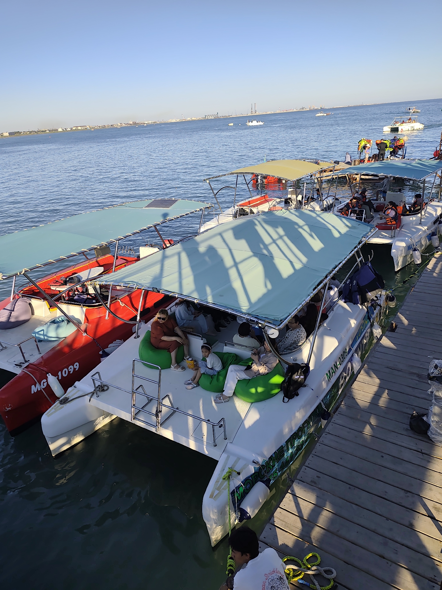 In the city of Unknown, Unknown, a high-angle, medium shot captures several catamarans docked at a wooden pier on a body of water under a clear sky. The catamarans, some red and white, others white, are filled with people. On the closest white catamaran, several individuals are seated on large green beanbag chairs, some interacting and others looking out at the water. A red catamaran with "MG 1099" visible on its side is docked alongside. More catamarans are visible further out in the water, some with canopies providing shade for passengers. In the background, a distant city skyline with buildings and industrial structures can be seen along the horizon. The water is calm and reflects the sunlight.