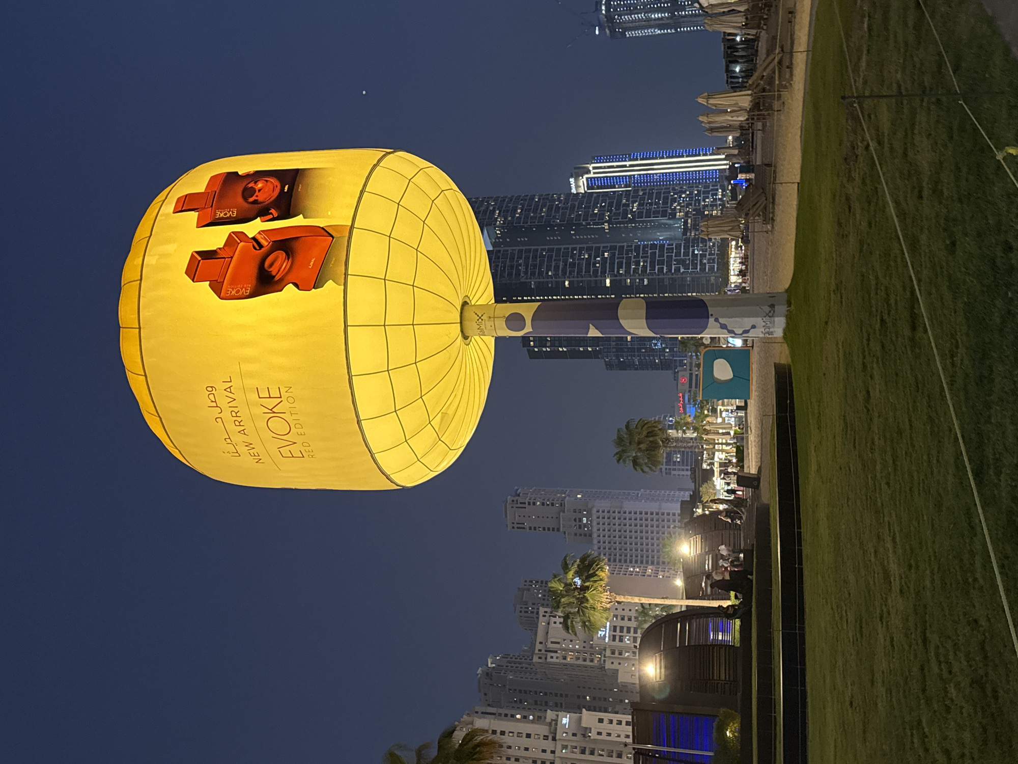 This is a nighttime, eye-level, medium-shot photograph of a large, illuminated yellow balloon advertising "EVOKE RED EDITION" perfume. The balloon is mounted on a tall, cylindrical pole with blue and white markings. In the background, a cityscape of Dubai, United Arab Emirates, is visible, with numerous skyscrapers illuminated against the dark blue night sky. The location appears to be a beachfront area, as evidenced by the sand, beach umbrellas, and lounge chairs. There is a grassy area in the foreground, and several people are visible in the middle ground, near some dark, curved structures. Two red perfume bottles are depicted on the balloon's surface. The overall scene suggests a vibrant, modern urban environment.