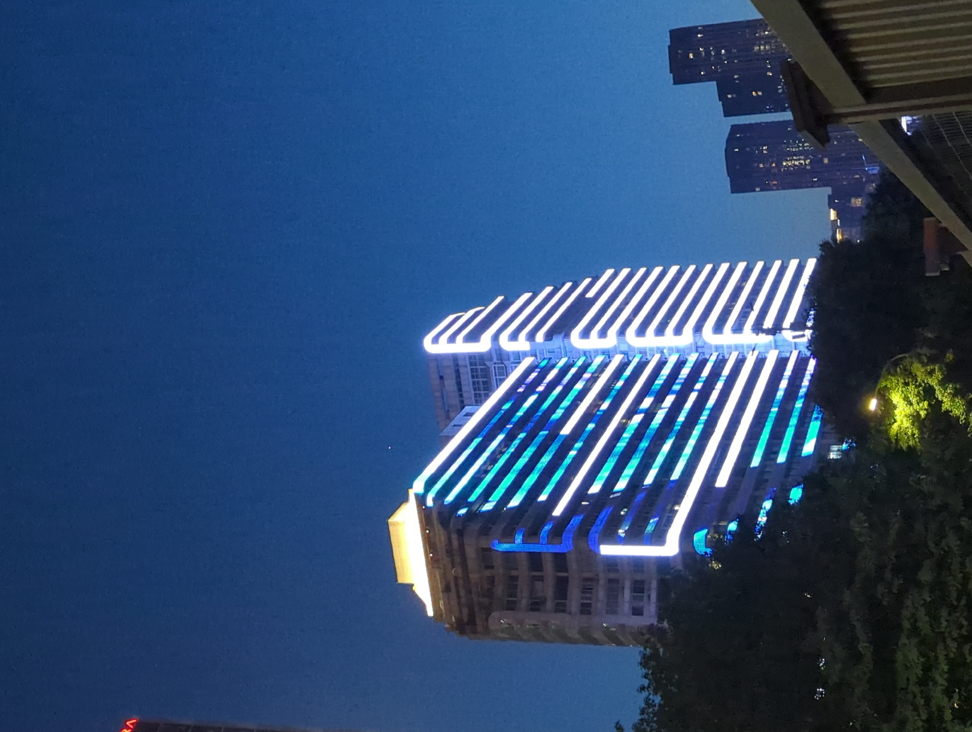 The image shows a night view of a modern cityscape in Yuzhong District, China. A tall, multi-story building is prominently featured, its facade adorned with a striking light display of alternating blue and white horizontal stripes. The building appears to have a unique, stepped design on its right side, with the lights accentuating this architectural feature. To the left of this building, another, older building with visible windows stands partially obscured by trees. Further in the background, several other skyscrapers with illuminated windows are visible against the dark blue night sky. The foreground is occupied by the dark silhouettes of trees and a portion of a wooden fence or railing. A streetlamp with a green-yellow glow is visible amidst the foliage on the lower right side of the image. The overall impression is one of urban development and nocturnal illumination.