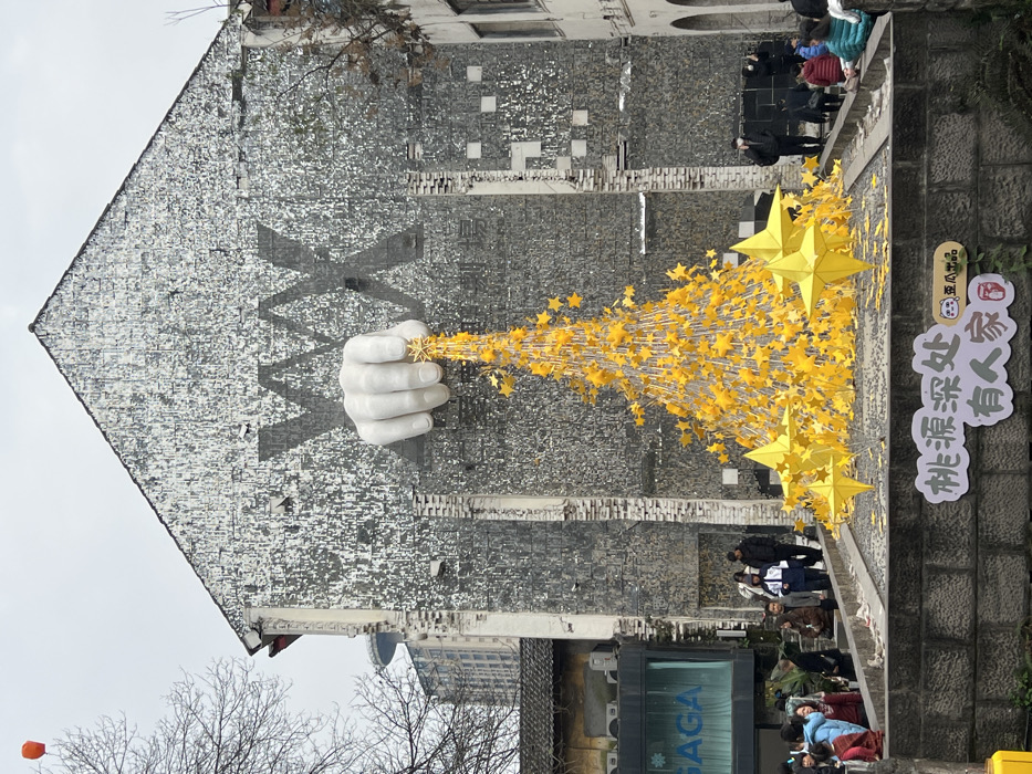 A building facade features a large mural of a hand seemingly holding a star, from which a stream of golden stars extends toward the ground. People are walking along the sidewalk near the base of the mural. A sign with Chinese characters is visible next to the stars, and a modern building with a glass storefront is partially visible in the lower frame. The sky above is overcast and there's a bare tree in the foreground.