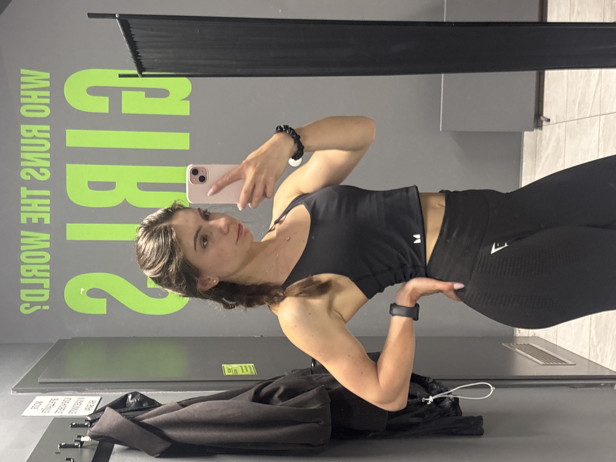A young woman is depicted taking a mirror selfie in what appears to be a gym or fitness studio in Varna, Bulgaria. She stands slightly to the left of the frame, looking directly into the smartphone she holds with her right hand. Her left hand rests on her hip in a posed manner.
The woman has dark brown hair, pulled back into a casual style, and a neutral expression. She is wearing a black athletic crop top with thin straps and black high-waisted seamless leggings. A small white logo, possibly an "M" or similar, is visible on her top, and another white logo, likely Gymshark's shark logo, is on her left hip on the leggings. She has a black scrunchie on her right wrist and a black fitness tracker or smartwatch on her left wrist. A delicate necklace with a small pendant adorns her neck.
The background features a grey wall with a large, bright green graphic reflected in the mirror. The visible text reads "GIRLS WHO RUN THE WORLD". Below this, dark grey or black ledges and surfaces are present. A black bag or jacket is visible on the floor in the lower left foreground. The lighting is bright and even, typical of an indoor public facility. The activity captures a moment of self-documentation within a fitness setting.