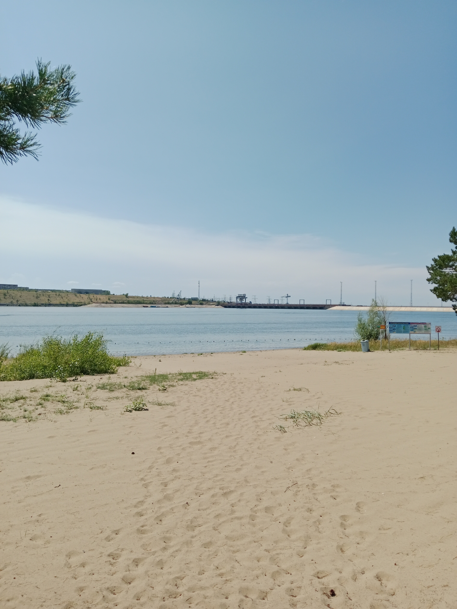 The image shows a sandy beach on a sunny day, with a body of water in the background. On the far side of the water, there is a large dam structure with several tall towers. In the foreground, there are footprints in the sand and some sparse vegetation. To the right of the image, there is a tall tree and a signpost with information and a red circle with a diagonal line, indicating prohibition. The sky is clear and blue. This appears to be a scene at the Bukhtarma Hydroelectric Power Station located near the village of Shulbinsk in Kazakhstan.
