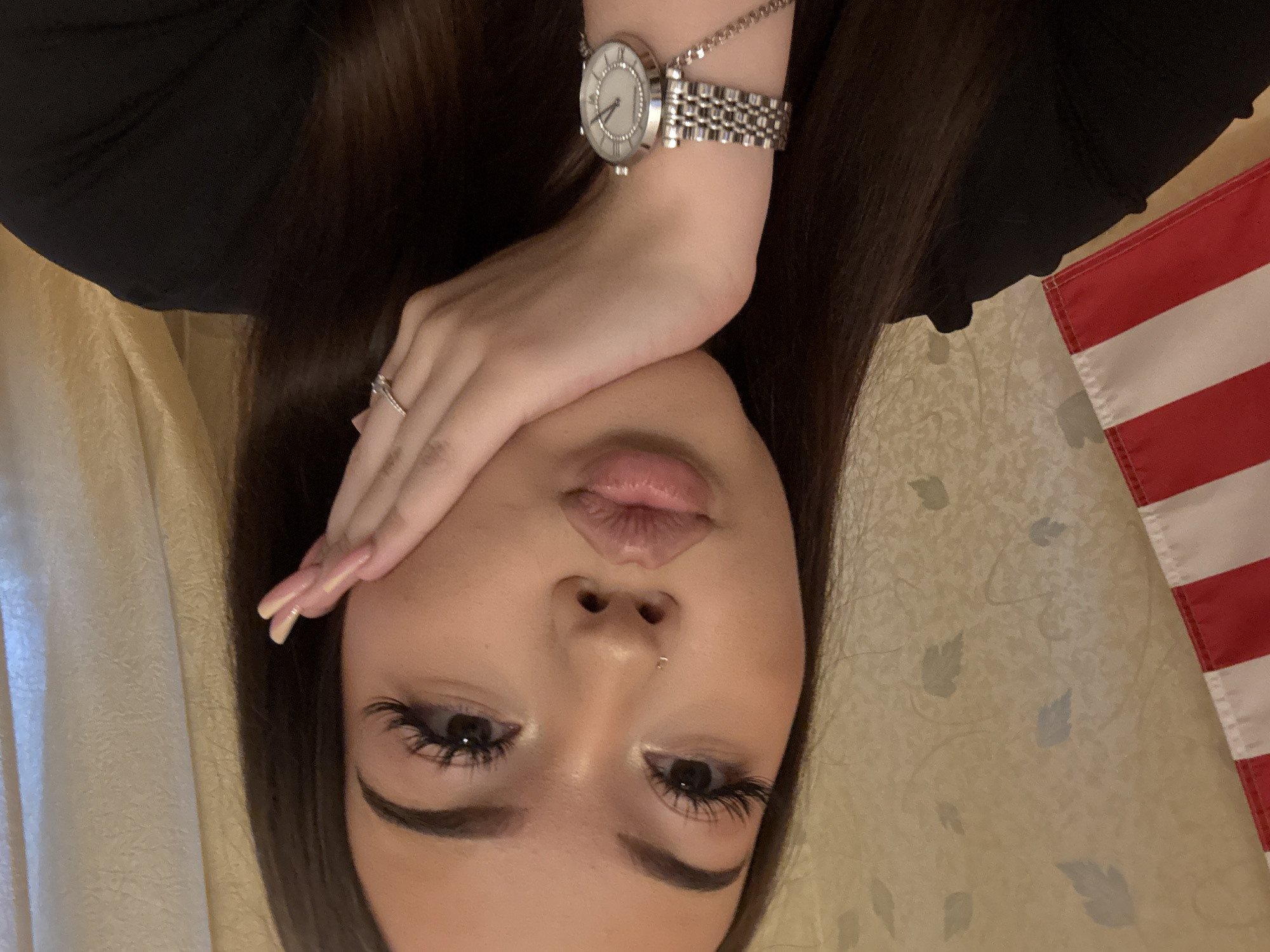A close-up portrait, oriented counter-clockwise, features a person, likely a young woman, lying down and looking towards the camera. She has long, dark brown hair and wears a black top. Her left hand, adorned with a silver watch and a silver ring, rests on her forehead, partially covering her eyes. Her nails are long and manicured with a light pink shade. Her facial features include full, slightly parted lips in a pout, prominent eyelashes (appearing to be false), well-defined eyebrows, and a small philtrum piercing.
The background consists of a light beige or cream-colored fabric, possibly a bedspread or curtain, with a subtle, lighter-toned leaf or vine pattern. On the right side of the frame, a section of a red and white striped flag, consistent with the design of the American flag, is visible. The lighting is soft and even, suggesting an indoor setting. The pose indicates a staged selfie. No specific time of day or weather can be determined from the image. The photo was taken in Naberezhnye Chelny, Russia.