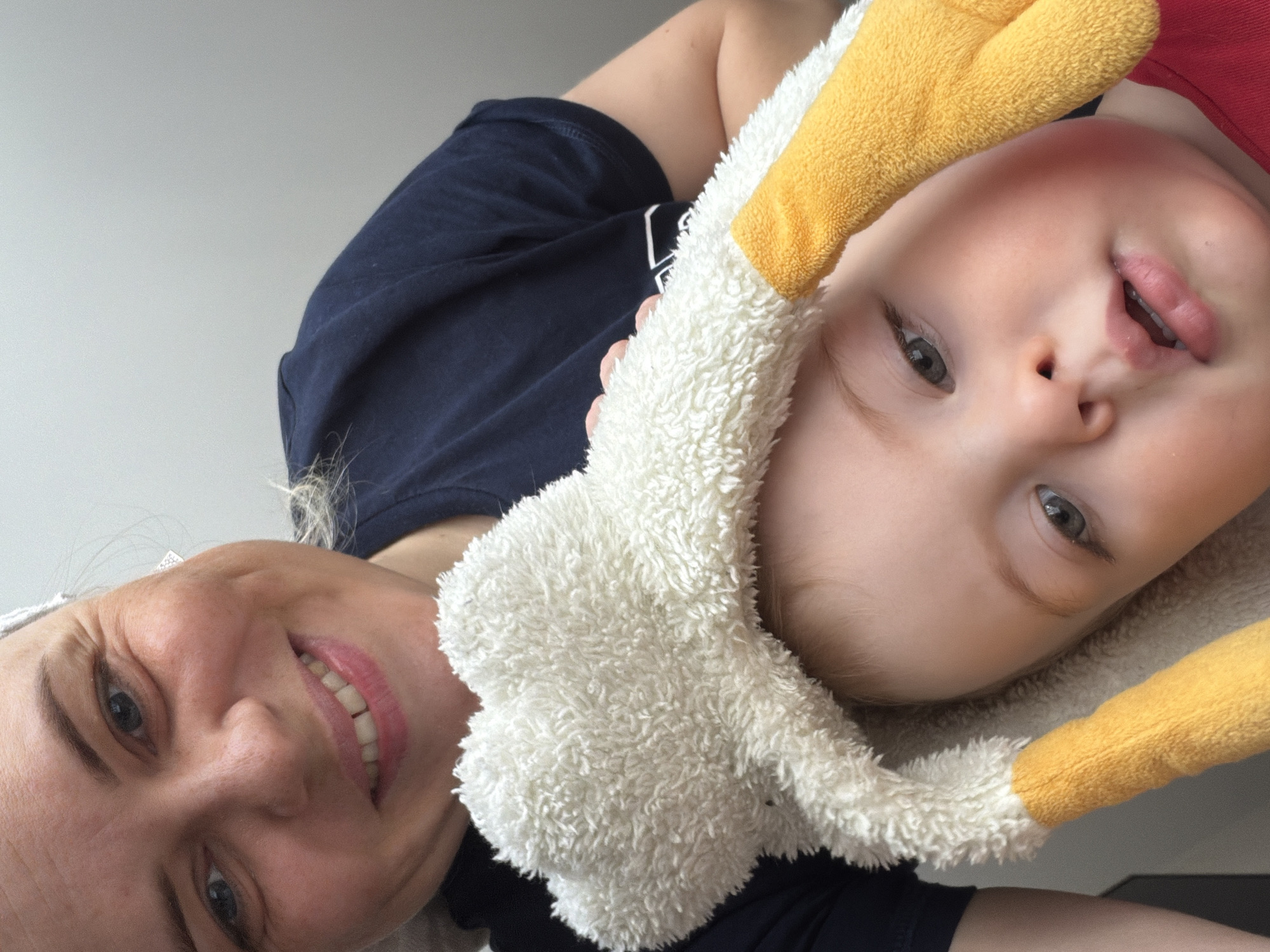 A mother and her baby are captured in a close-up, tilted selfie. The mother, on the left, smiles broadly, her face close to the camera. To her right, the baby looks at the camera with an open mouth. The baby wears a stuffed animal on its head, white with yellow accents. Both the mother and baby wear dark-colored clothing. The background appears to be a neutral-colored wall. The location is unknown and does not seem to be Votkinsk, Russia.