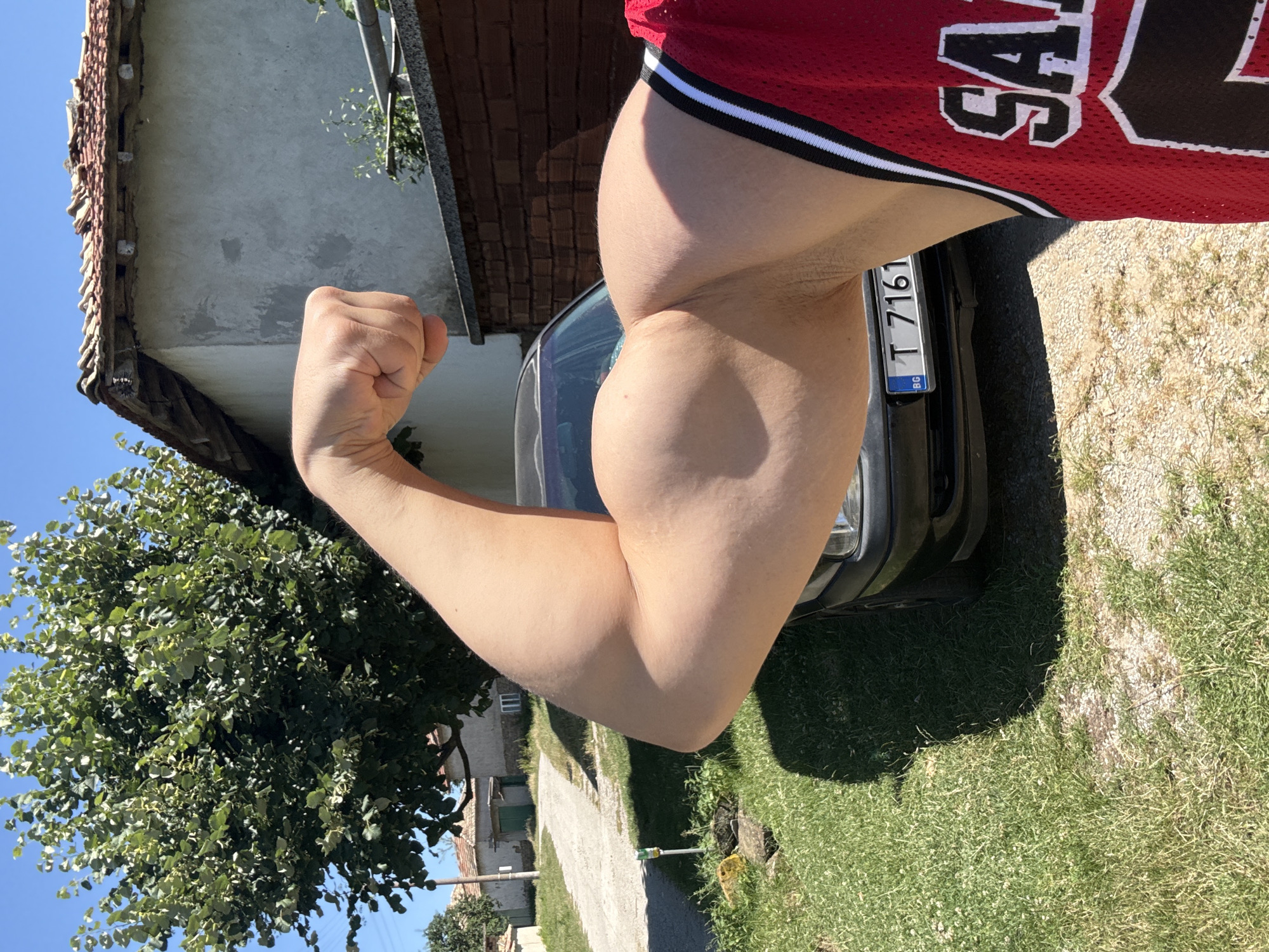 The image depicts a close-up of a man's flexed bicep, with his fist clenched and held aloft. He is wearing a red athletic jersey with white trim. Behind him, a black car is parked, with a license plate visible that reads "BG T 7161". The scene appears to be outdoors, with a rural setting evident. A dirt road and green grass are present, along with a house with a tiled roof and a large tree. The lighting suggests a sunny day. The car and surrounding buildings suggest a location in a village or town, possibly Konop, Bulgaria, given the license plate format.