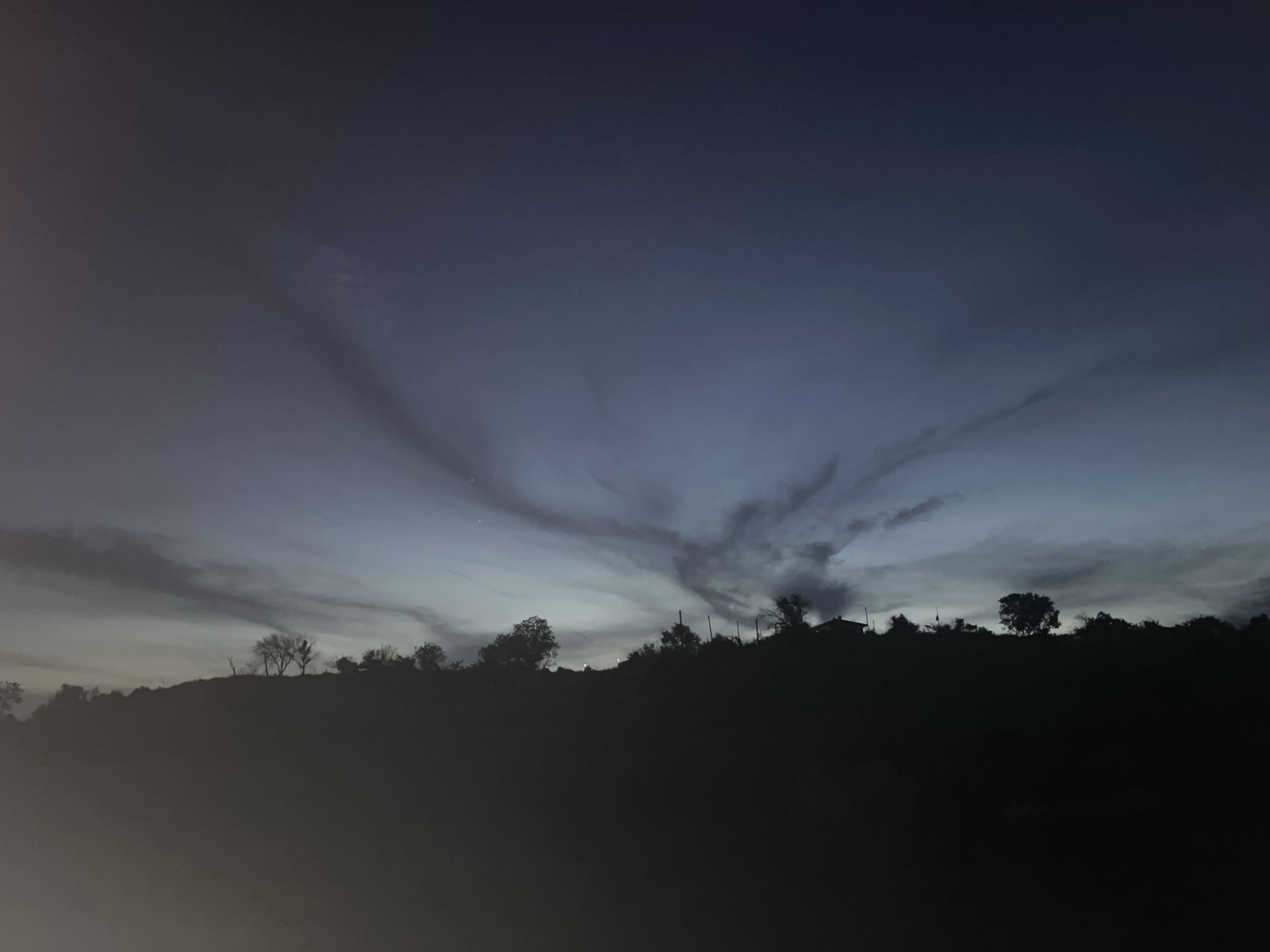The image depicts a dark, silhouetted landscape at dusk or dawn. A prominent hill or ridge dominates the lower half of the frame, its contours defined by the faint light from the sky. The top of the ridge is dotted with the dark shapes of trees, some appearing bare and skeletal, while others are fuller. A few indistinct structures, possibly houses or outbuildings, are visible on the ridge, silhouetted against the sky.
The sky above is a gradient of deep blues and grays, with wispy, elongated clouds stretching across the frame. These clouds catch the fading light, creating a sense of depth and texture. The overall mood is somber and quiet. The image does not contain any identifiable people or specific landmarks that would allow for pinpointing a location within the city of Varna, Bulgaria. The scene is generic to any hilly, vegetated area observed during twilight.