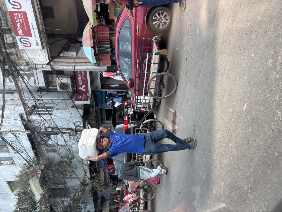 A man in a blue shirt and jeans carries a large white package on his head. He walks on a paved street. Behind him are bicycles and cycle rickshaws. A red car is parked behind a rickshaw. Buildings with tangled wires are on the left. One building has signs in a foreign language. A few other people are visible in the background.