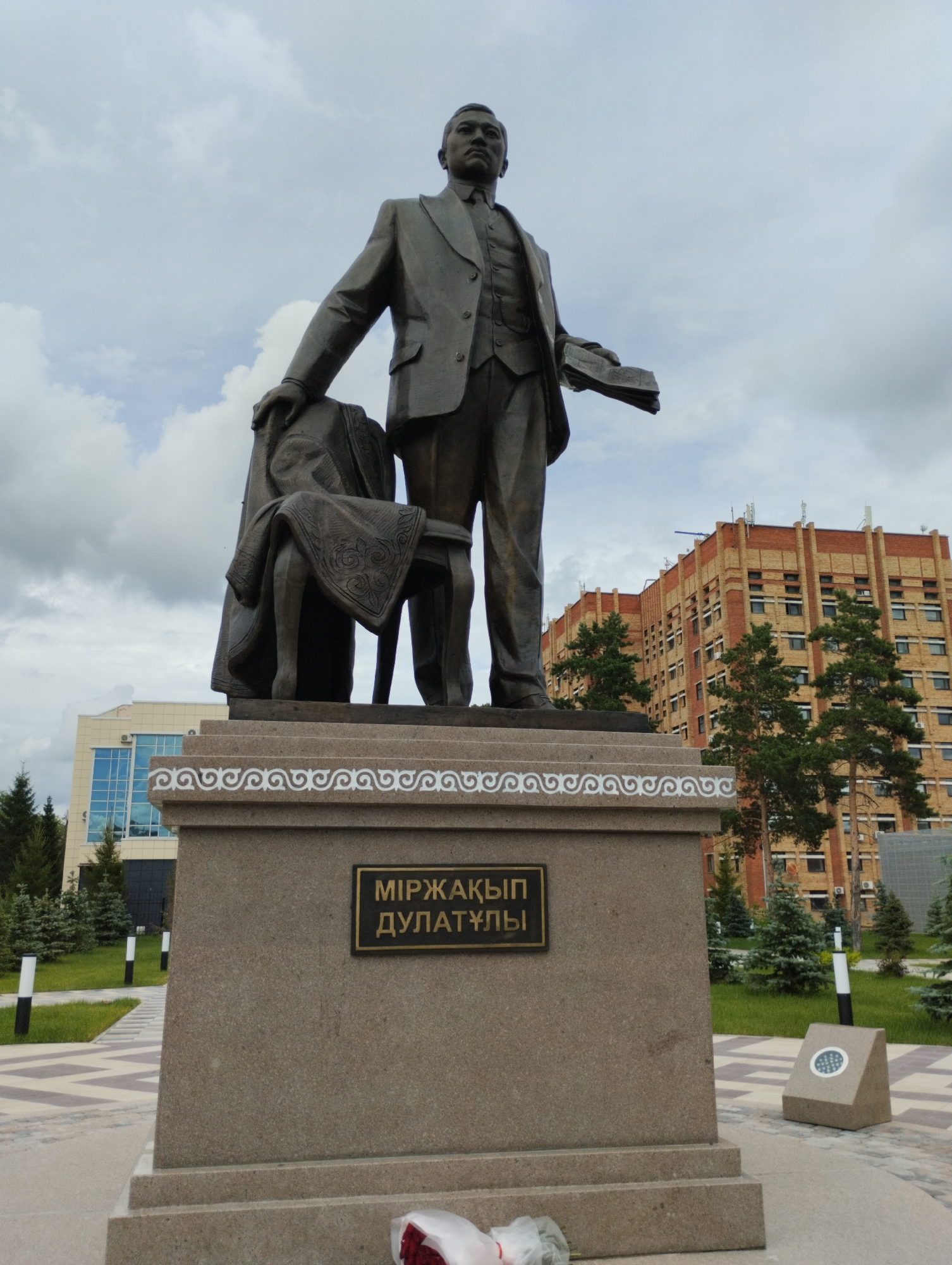 The image depicts a bronze statue of a man in a suit, standing next to a draped chair. The statue is mounted on a granite pedestal with an inscribed plaque that reads "Мiржакып Дулатулы". The background shows a modern, multi-story brick building with numerous windows. To the left of the building, there is a blue-windowed structure. Several evergreen trees are visible around the statue and the buildings. The scene appears to be an outdoor public space, possibly a park or square, with paved pathways and lampposts. The sky is cloudy. The statue and its pedestal are centrally located in the foreground. The specific location is identified as the Mirzakhip Dulati monument in Kostanay, Kazakhstan.