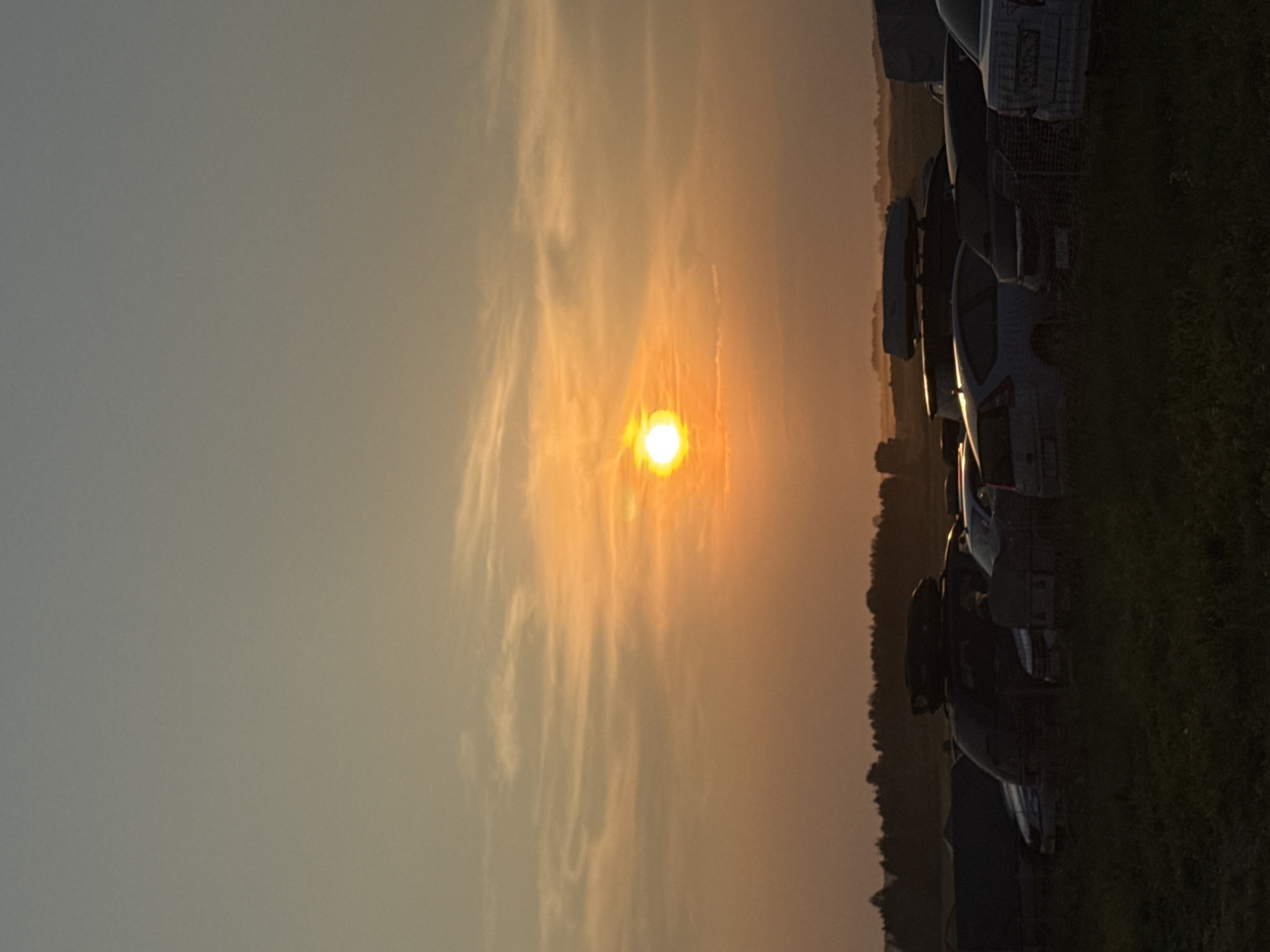 The image depicts an outdoor scene at dusk, likely in a rural or suburban area, given the presence of a treeline and open fields. The sky is a gradient of soft oranges and grays, with the sun, a bright orb, low on the horizon, casting a warm glow. A line of parked vehicles, including cars and what appear to be vans or SUVs, are visible in the foreground and midground, some covered with tarps or with roof cargo carriers. The vehicles are parked on a grassy surface. In the distance, a silhouetted treeline marks the horizon. The overall atmosphere is tranquil and serene, characteristic of the transition from day to night. No people are visible in the frame.