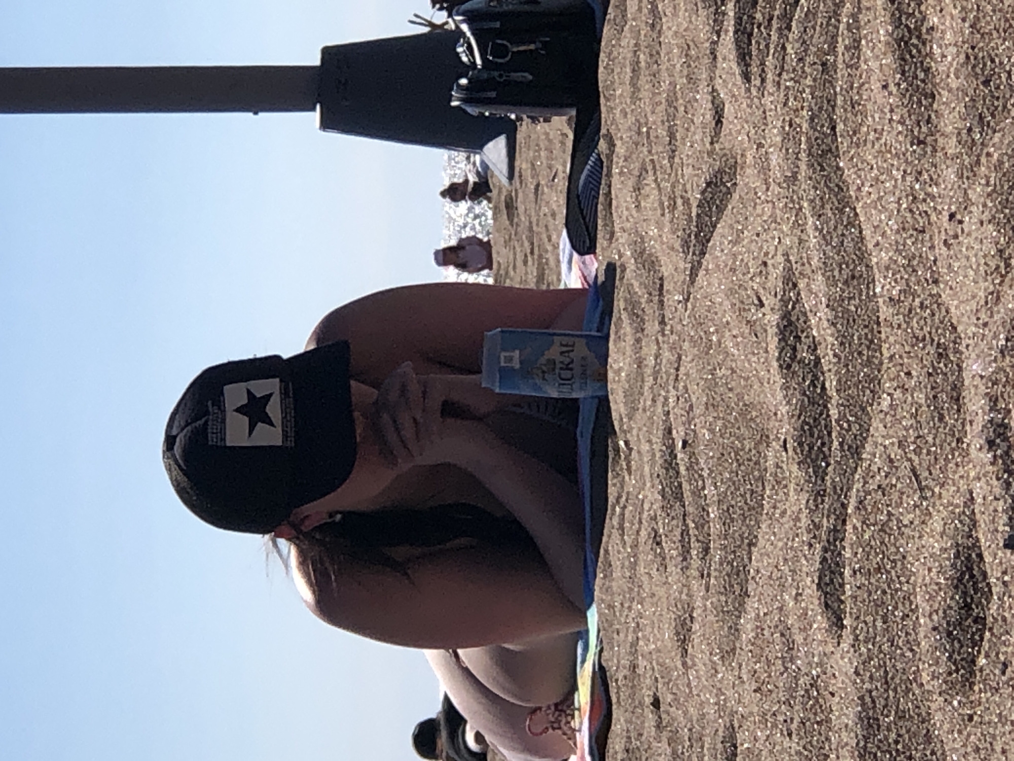 The image depicts a beach scene in Solnechnoye, Russia, under clear blue skies. In the foreground and mid-ground, an individual is positioned prone on a multi-colored striped beach towel (featuring blue, red, and yellow hues). The individual wears a black baseball cap with a white rectangular patch on the front, bearing a black five-pointed star. Their hands are clasped in front of them, and long dark hair is visible beneath the cap. An earring is discernible on their left ear. To the immediate right of the individual, a blue and white beverage can with Cyrillic text, reading "ДЮСКАЕ" (Dyuske), rests on the sand. Further to the right, a black bag or purse is placed on the sand, next to what appears to be dark-colored footwear. A dark, thick, vertical pillar-like structure rises on the far right of the frame. The foreground consists of light brown sand with distinct ripple patterns. In the background, beyond the immediate beach area, a sparkling body of water (likely the Gulf of Finland or a similar large body of water) is visible under bright sunlight. Several other indistinct individuals are present in the mid-ground and background, scattered along the shoreline or in the water.