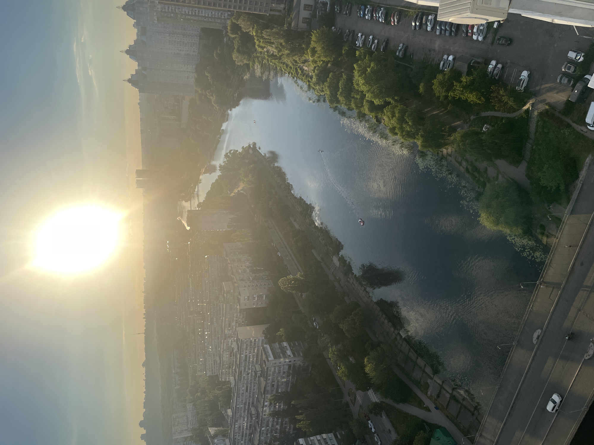 This is an aerial view of the Dnipro River in Kyiv, Ukraine, at sunset. The sun is low on the horizon, casting a warm, golden light across the scene. Tall residential buildings line the riverbanks, interspersed with trees and parks. Several cars are visible on the road and in a parking lot along the river. Two small boats are on the water, and one person can be seen walking on a bridge in the foreground. The overall atmosphere is peaceful and serene.
