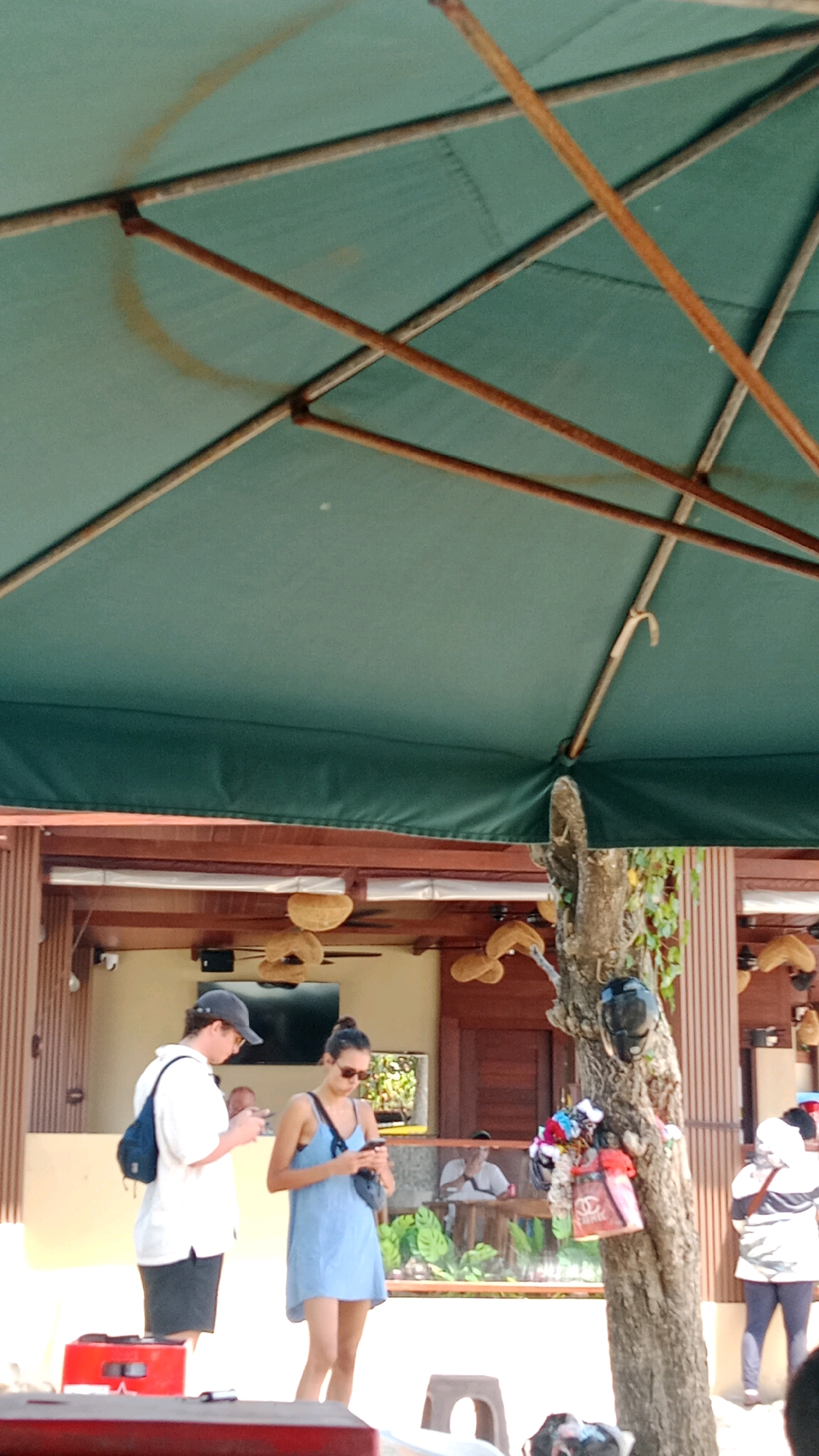 An outdoor scene under a large green umbrella in Kuta, Indonesia. A man in a white shirt and dark shorts, wearing a cap and a blue backpack, stands with a phone in his hand. Beside him, a woman in a light blue dress and sunglasses also holds a phone. To the right, a tree trunk is decorated with colorful accessories. In the background, a building with wooden accents and hanging straw decorations can be seen, along with other people seated at tables. The overall atmosphere suggests a casual, possibly beachfront, establishment.
