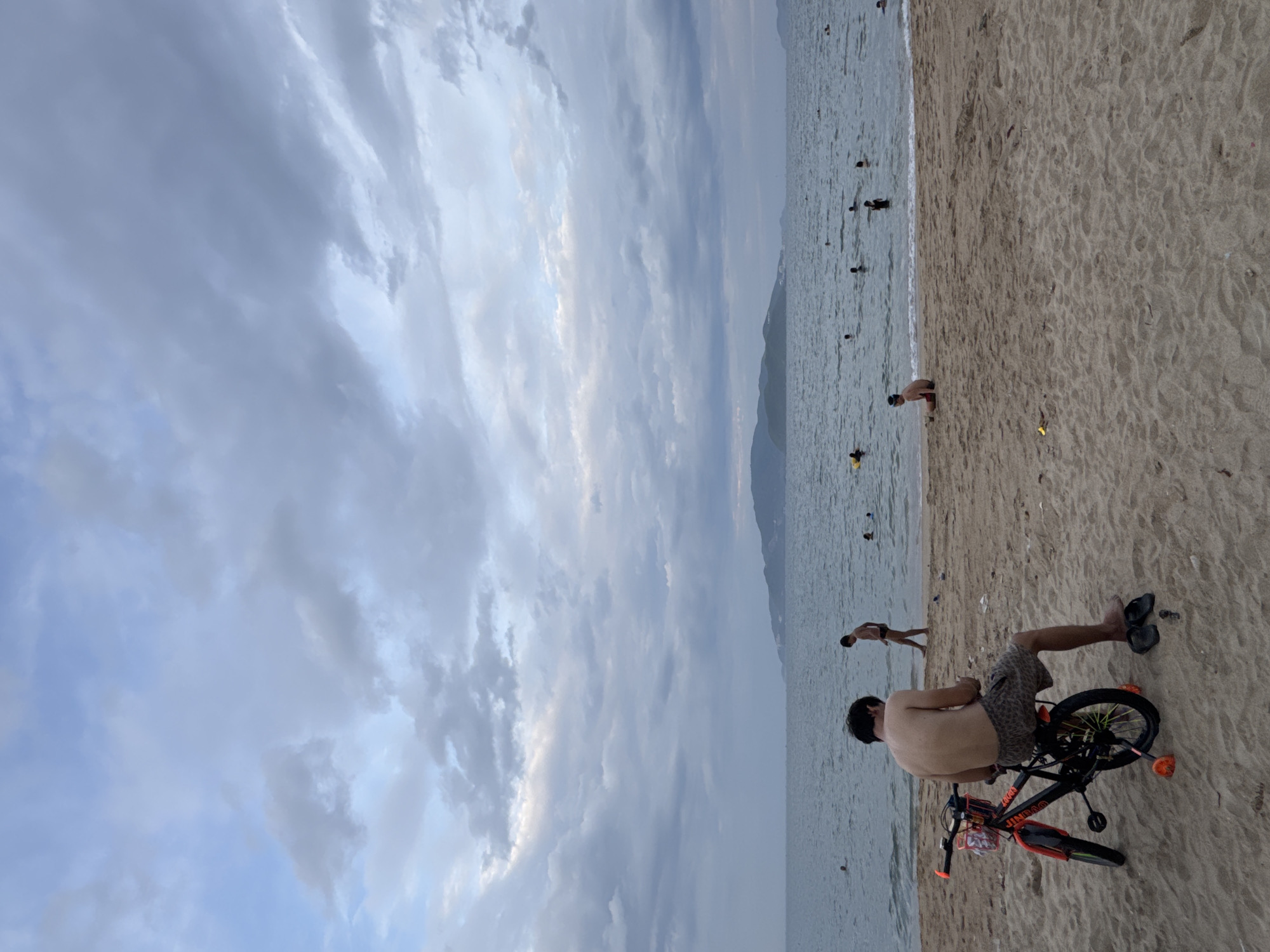 The image depicts a beach scene in Nha Trang, Vietnam, under a cloudy sky. In the foreground, a young man sits on a bicycle, facing away from the camera, appearing to be using his phone. The bicycle is black with orange accents and has training wheels. The sand is light brown, and a pair of black flip-flops rests beside the bicycle. In the middle ground, several people are visible on the beach and in the shallow water, with one individual walking towards the sea. The sea itself is a muted blue-grey, stretching out to the horizon where a low, green island chain is visible. The sky is filled with a dense layer of grey and white clouds, with hints of lighter sky peeking through.