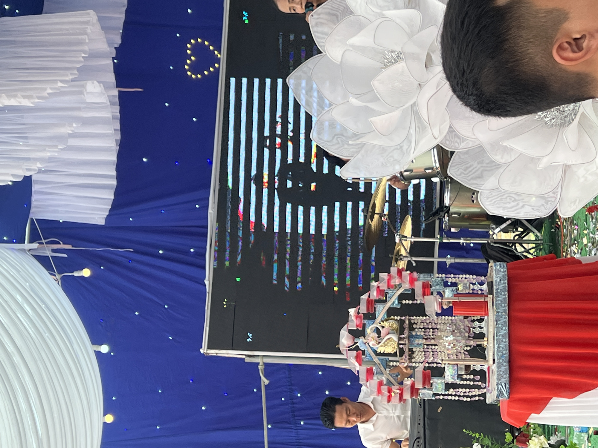 A horizontal indoor shot shows decorations and people at a ceremony in Tay Son Commune, Vietnam. A blue backdrop with twinkling lights forms the background. Decorations include large white floral arrangements, white draped elements, and an elaborate beaded and bejeweled structure with red accents and golden swans. A digital screen displaying vertical lines and a silhouette of a person stands prominently. Two men are visible, one in a white shirt and one in the upper right corner with short black hair. The man in the white shirt is positioned near the beaded structure, while the other is partially obscured. The scene suggests a formal event with a blend of traditional and modern elements.