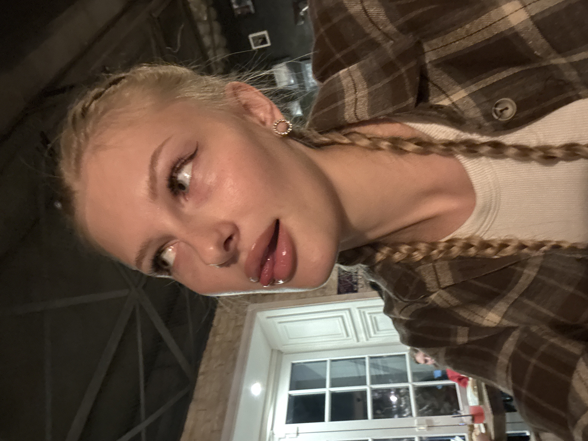 The image is a close-up selfie of a young blonde woman with braided hair, wearing a plaid shirt and a white tank top. She has on makeup, including eyeliner and lip gloss. Her tongue is sticking out slightly, and she is looking upwards. She is wearing a small circular earring. In the background, there are windows, a table with drinks, and other people. The scene appears to be indoors, possibly a restaurant or bar. There is no discernible information in the image to suggest a location in the city of Perm, Russia.