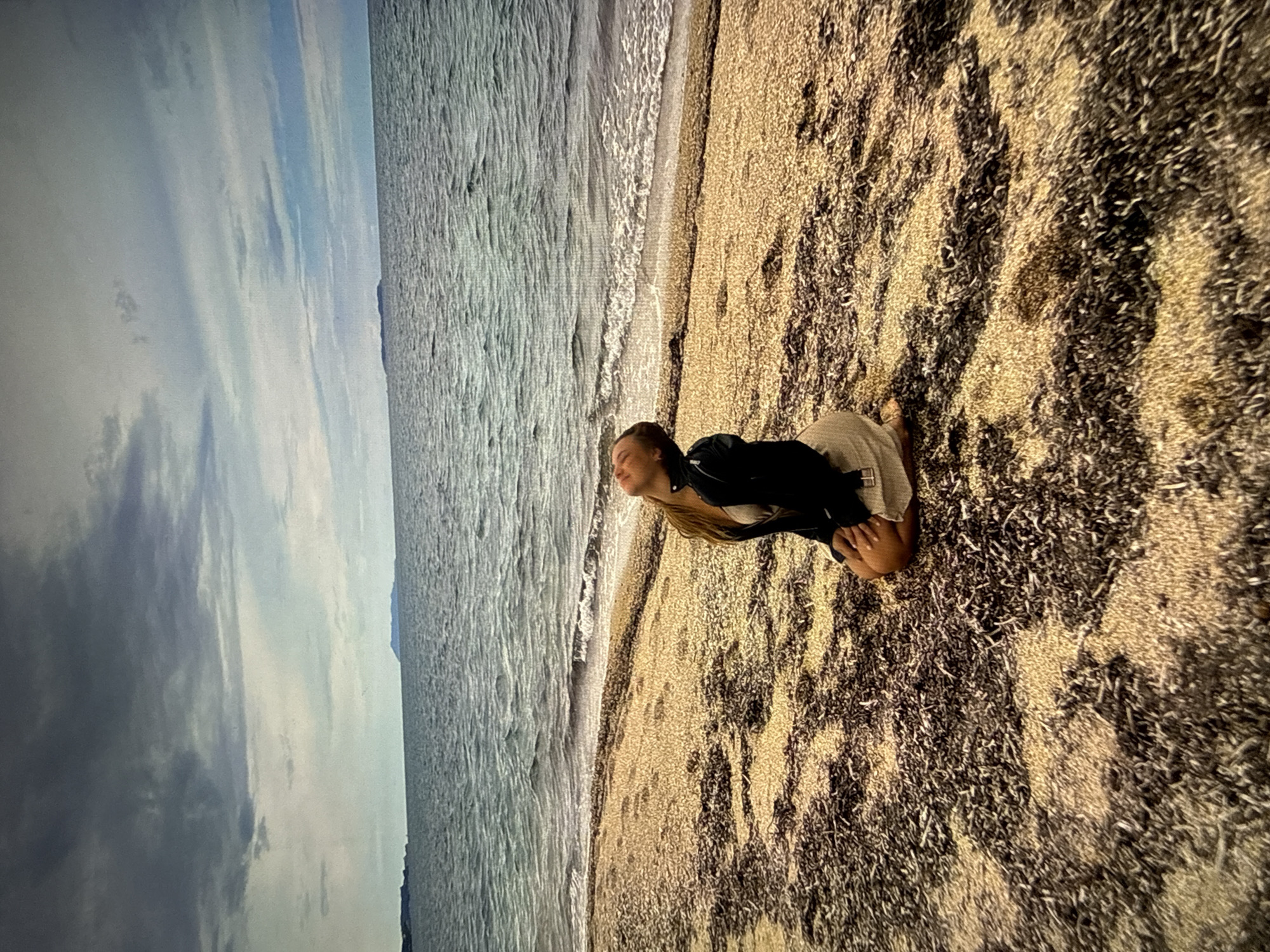 A woman kneels on a beach. Her long blonde hair is pulled back, and she wears a black jacket over a light-colored top and bottoms. The beach has a sandy texture with dark patches. The ocean is visible in the background, with waves meeting the shore. The sky is overcast with grey and blue clouds. The image is rotated ninety degrees counter-clockwise. This scene does not appear to be in Sofia, Bulgaria, as Sofia is an inland city. The scene is more suggestive of a coastal location.