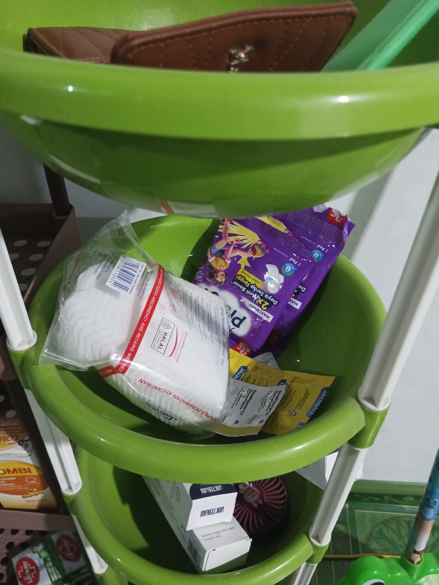 The image shows a close-up view of a three-tiered green plastic shelving unit containing various items. The top shelf holds a brown quilted wallet. The middle shelf contains a package of white cotton pads and a purple package of maxi pads with cartoon characters. Beneath the maxi pads are two small yellow packets. The bottom shelf contains a white box labeled "DOKTERIGI", a pink fan-like object, and a partially visible yellow and orange box. The overall environment appears to be indoors, possibly a home or a retail setting, with a hint of a wall and floor visible. The lighting is dim and the image is slightly blurry. There are no people or actions depicted in the image. The location is stated as Pohuwato, Indonesia.