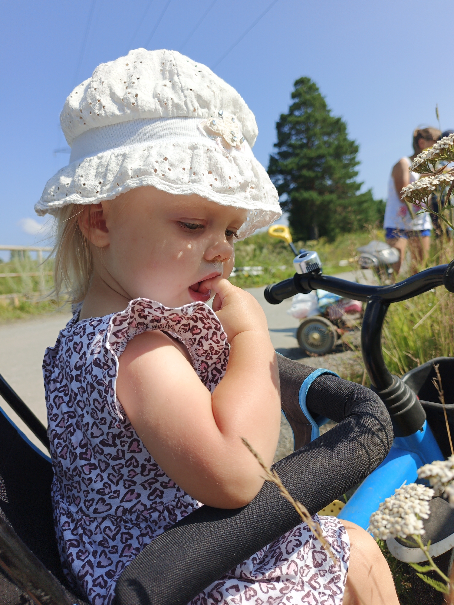 The image shows a young child wearing a white sun hat and a patterned dress, sitting in a stroller with a blue frame. The child has their finger in their mouth and is looking down. In the background, a woman in a white tank top and blue shorts is visible, as well as a tree and a clear blue sky. The scene appears to be outdoors, possibly in a park or rural area. The image does not provide any information about the city of Urtiushka, Russia.