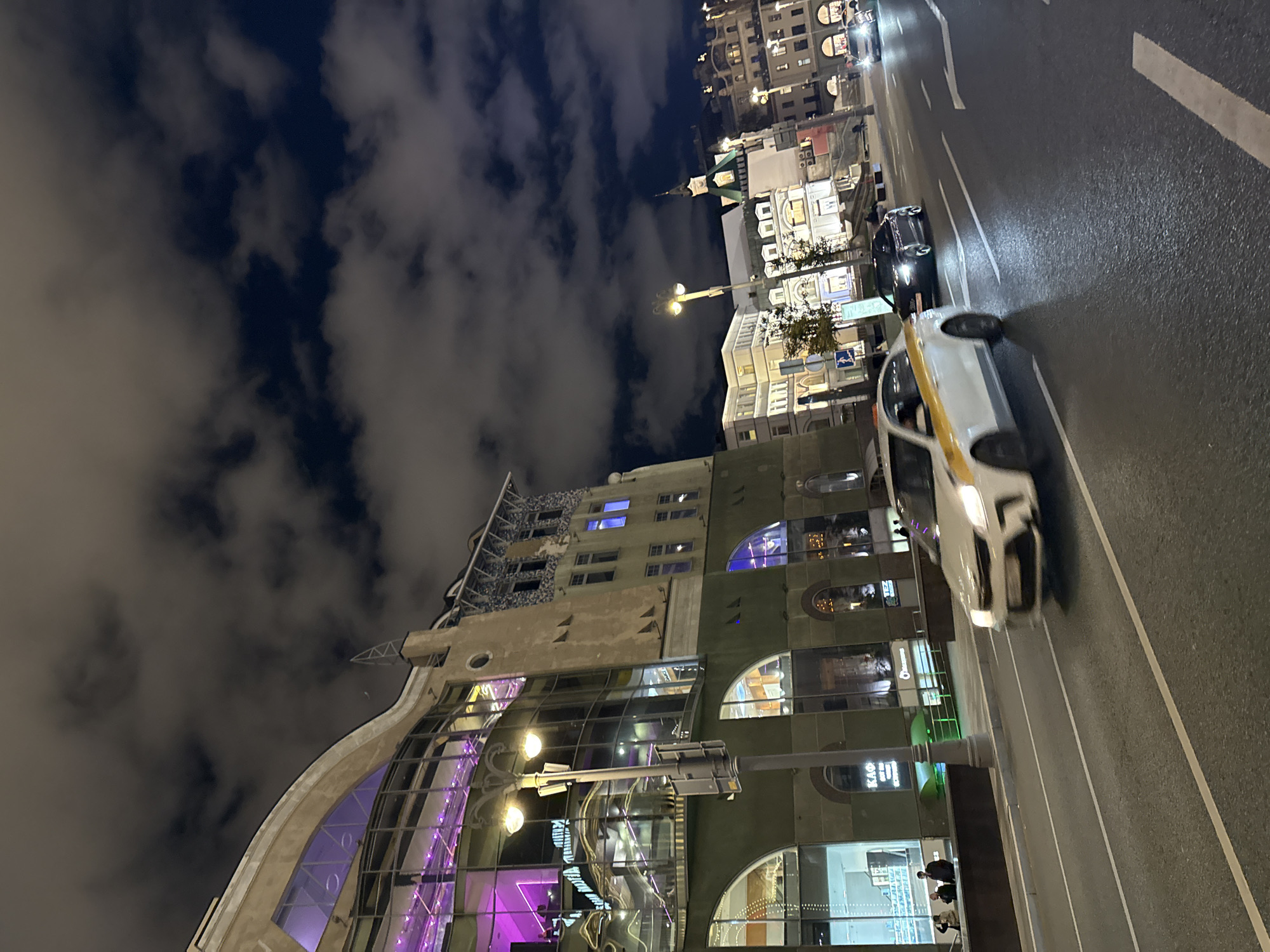 The image shows a night-time street scene in Moscow, Russia, likely in a busy urban area. A white taxi with yellow accents is in motion on the right side of the road, blurring due to its speed. To the left of the taxi, a dark-colored sedan is also visible on the road.
Dominating the left side of the frame is a multi-story building with a unique architectural style. The lower floors feature large arched windows, with some illuminated from within. The upper portion of the building has a beige facade with intricate decorative elements and several windows. This building appears to be a commercial establishment, possibly a restaurant or a club, as indicated by the vibrant purple lighting emanating from some of the upper windows. A streetlamp with two illuminated globes stands in front of this building.
In the background, across the street, are other buildings with illuminated windows, suggesting a lively urban environment. The sky is dark with scattered, fluffy clouds, illuminated by ambient city lights. The street is paved and marked with white lane dividers. There are a few people visible on the sidewalk in front of the building on the left, and other vehicles and pedestrians are present further down the street in the background.