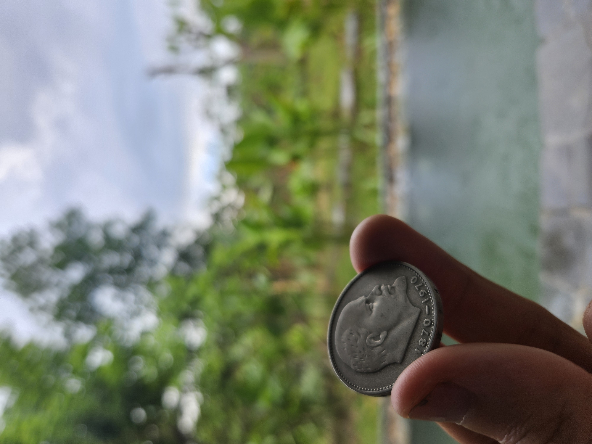 A hand is holding a silver coin depicting a profile of Vladimir Lenin, with the dates "1870-1970" engraved below. The coin is held in the foreground, with a blurred background of lush green foliage and a body of water. The sky above is overcast. The image does not provide any specific visual cues that can identify the location as Đà Nẵng, Vietnam.