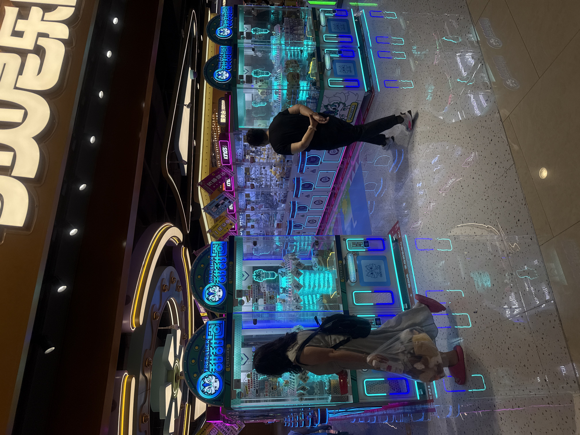The image depicts the interior of a brightly lit arcade, likely a gaming center, in Qinhuangdao, China. The scene is dominated by rows of colorful arcade machines, prominently featuring claw machines with plush toys visible inside. Neon lights, primarily in shades of blue and pink, illuminate the machines and reflect off the polished floor, creating a vibrant and somewhat futuristic atmosphere. Two individuals are visible. On the left, a woman walks away from the viewer, carrying a bag with stuffed animals, wearing a light-colored dress and pink slippers. On the right, a man, seen from the back, walks towards the arcade machines, dressed in a black t-shirt and dark pants. The overall impression is a lively entertainment venue with a focus on gaming and prizes.