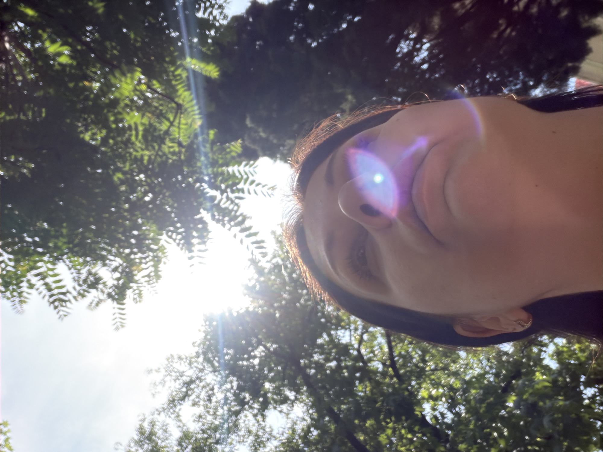 The image captures a low-angle perspective of an individual looking upwards, framed by sunlit tree canopies against a bright sky, located in the city of Sochi, Russia. The lower half of the frame features the upper face of a person, likely female, whose dark hair cascades around her head. Essential facial features visible include her nose, mouth, chin, and part of her left eye, with a small, reflective earring in her right earlobe. Intense sunlight from above creates prominent lens flares, casting a bright purple and white glow across her nose and mouth, and a linear streak on the upper right. The environment consists of dense green foliage from multiple trees, some with fine leaves and others with broader, darker forms, backlit by a brilliant sky that transitions from bright white near the sun to a pale blue. The overall scene depicts a serene outdoor setting under clear, sunny conditions.