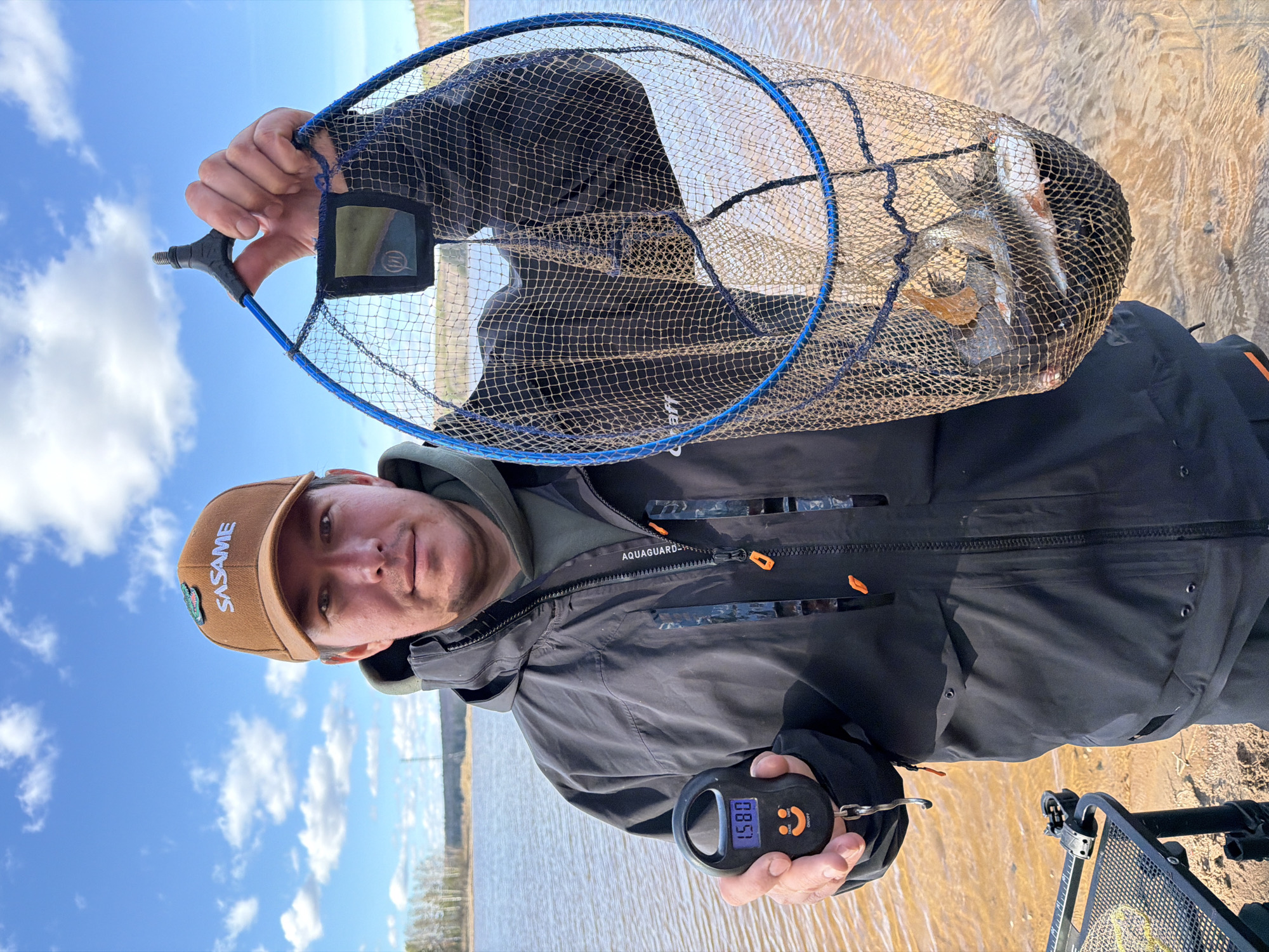 A man in a brown hat and black jacket stands outdoors near a body of water under a partially cloudy sky. He holds a blue fishing net containing several fish over his head with his right hand, and a black digital scale in his left hand showing a weight of 1.580. The jacket bears the brand "AQUAGUARD." The hat has "SASAME" embroidered on it. There is a metal object on the ground nearby, possibly a fishing chair or equipment. Based on the horizon and landscape, the image appears to have been taken near Chaikovsky, Russia.
