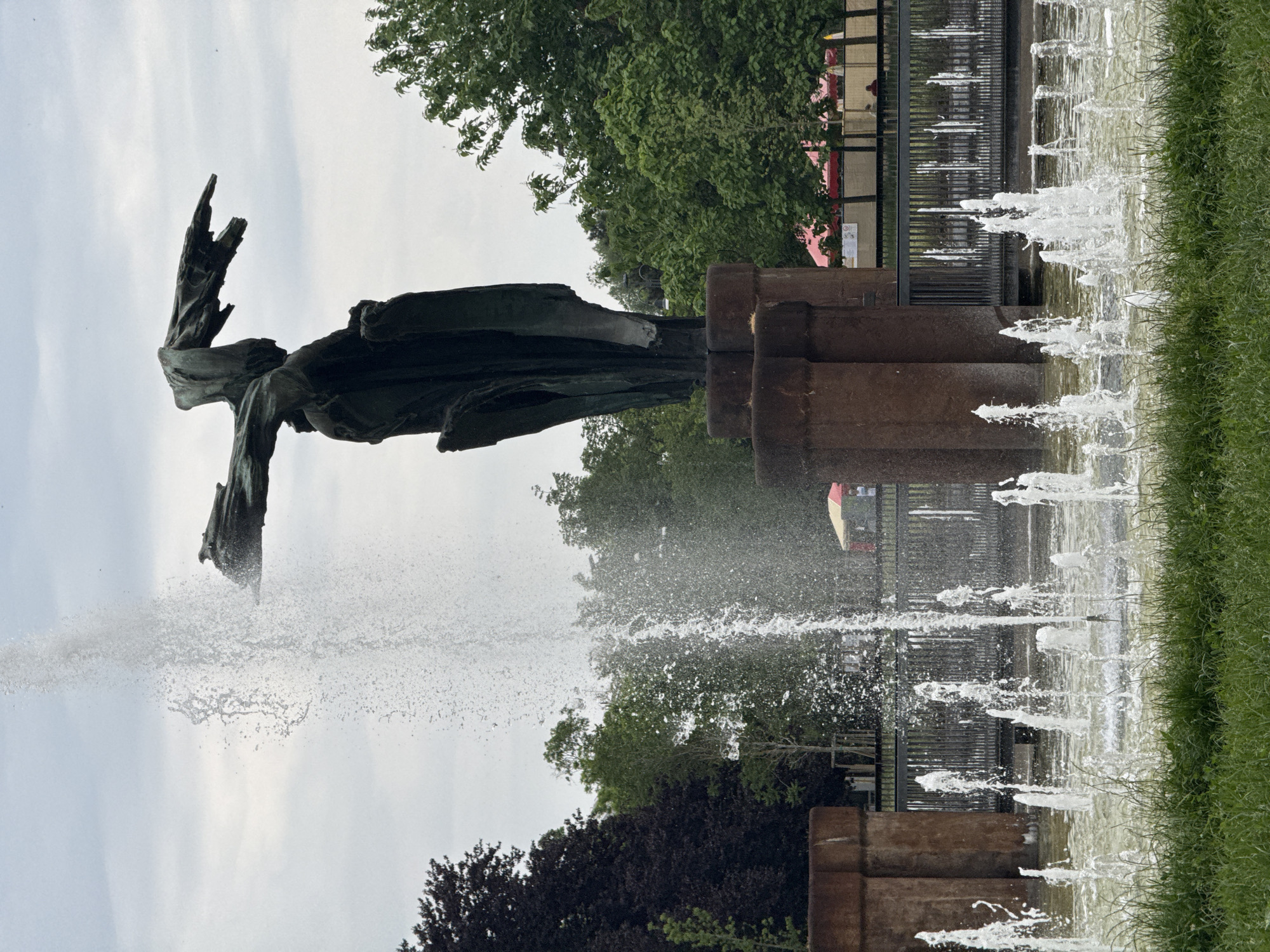 This image, when viewed upright, captures an outdoor fountain scene in Milan, Italy. A large, dark, stylized sculpture, possibly bronze, stands prominently on the left. The figure features outstretched limbs or wings, contributing to a dynamic and abstract form. Water vigorously sprays from multiple points: directly from the base of the sculpture and from a long, rectangular pool that extends horizontally across the frame. These water jets create numerous vertical streams and a misty spray around the central figure.
Along the right edge of the fountain pool, a strip of vibrant green grass is visible. The background is dominated by lush green deciduous trees with full foliage, suggesting a park setting. Further back, beyond a dark metal fence, hints of distant buildings and indistinct figures, some wearing red clothing, can be seen, indicating a public area. The sky above is bright and largely overcast, suggesting a daytime scene, likely late morning or afternoon. No discernible text is visible in the image.