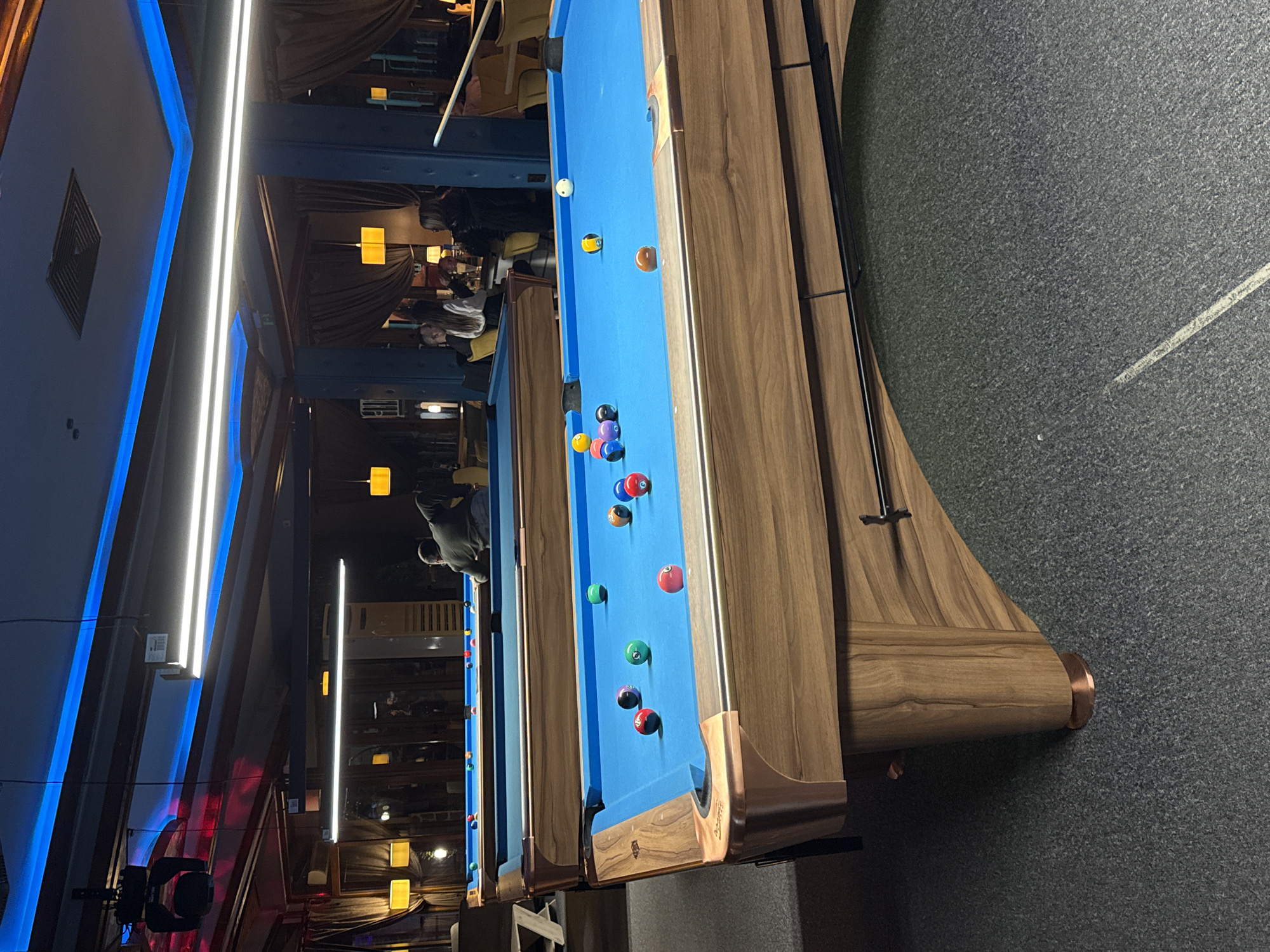 The image depicts a billiards hall with a row of pool tables covered in blue felt. Several billiard balls are scattered on the tables. In the background, blurred figures of people are visible in a dimly lit seating area. The tables have wooden frames. The flooring is dark gray, and the walls are partly visible in the background, showing architectural details with neon lighting. There is a high probability this location is in Plovdiv, Bulgaria, as that was where it was taken.