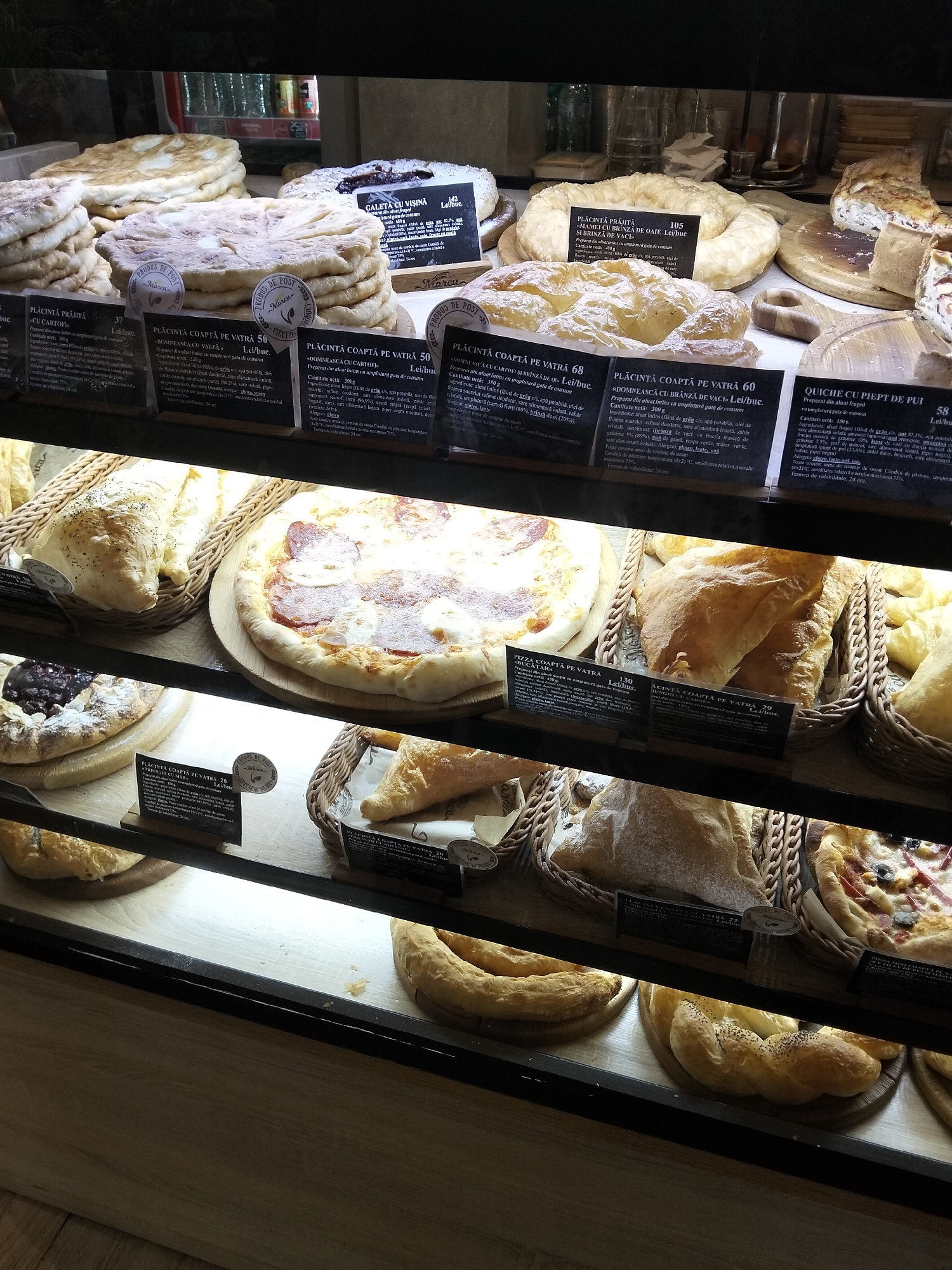 This media file captures a close-up view of baked goods displayed in a bakery case, likely in Peresecina, Moldova. The primary subjects are various types of bread, pies, and pastries arranged on shelves. The scene is an indoor setting, a bakery, illuminated by artificial light that highlights the golden-brown crusts and dusting of flour on the items.
The products include large, round flatbreads, some dusted with sugar, and other flaky pastries. There are also pizzas visible, one topped with cheese and salami. A tart with a dark berry filling is also present. Many of the items have small, circular labels or tags attached, indicating their names and possibly prices, written in Romanian.
The activity taking place is the display and presumably sale of baked goods. The arrangement suggests a retail environment where customers can select their purchases. The lighting creates a warm and inviting atmosphere, typical of a bakery.
Notable details include the visible text on the labels. For instance, one label reads "PLA CIN TĂ COAPTĂ PE VATRĂ" which translates to "Pie baked on a hearth". Another label states "QUICHÉ CU PIEPT DE PUI" meaning "Chicken breast quiche". There are also labels that mention "LEI/buc." indicating the currency. The overall impression is of a well-stocked bakery offering a variety of traditional and modern baked goods. The time of day is not discernible, nor is the weather outside, as the focus is solely on the interior display. No people are visible in the image.