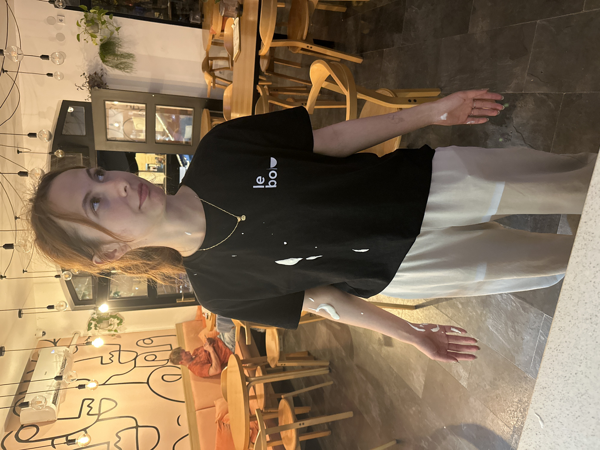 A young woman with light reddish-brown hair is standing indoors, facing left and looking upwards. She wears a black t-shirt with white lettering and white pants. Her hands are outstretched with small amounts of white substance smeared on them. She is also wearing a gold necklace. The background includes a cafe or restaurant setting with wooden chairs and tables, and decorative light fixtures. On the left is a pink wall with black graphic designs. The location is Novi Sad, Serbia, discernible by the "le bol" logo on the t-shirt, which is a cafe name located in Novi Sad, Serbia. A man is sitting on a couch in the background.