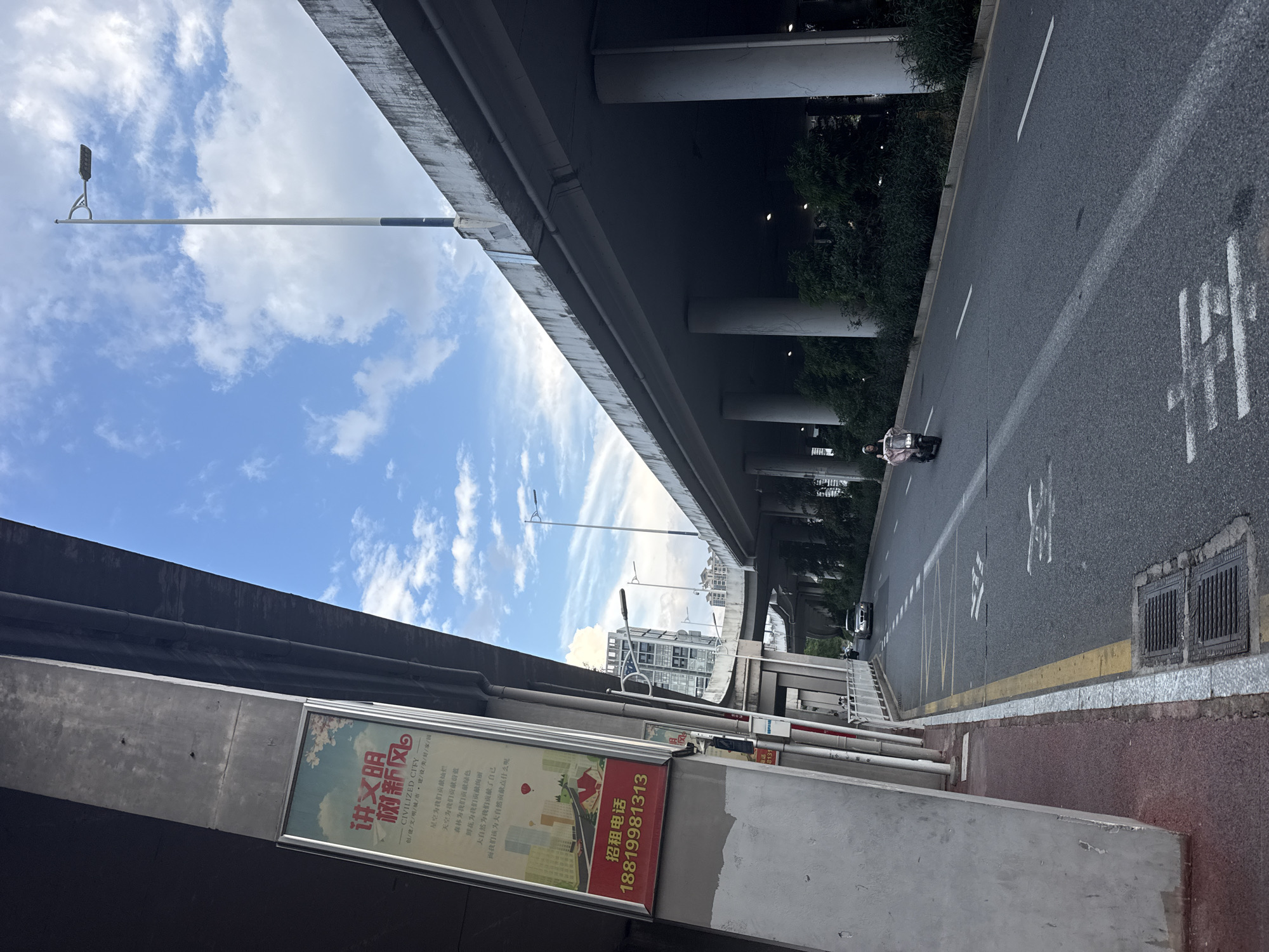 An outdoor medium shot shows an overpass in Huizhou, China. The overpass is made of concrete and is supported by large pillars. A street runs beneath the overpass, with a motorcycle rider heading away from the viewer. A billboard advertising "CIVILIZED CITY" is attached to one of the pillars. The sky is blue with scattered clouds.