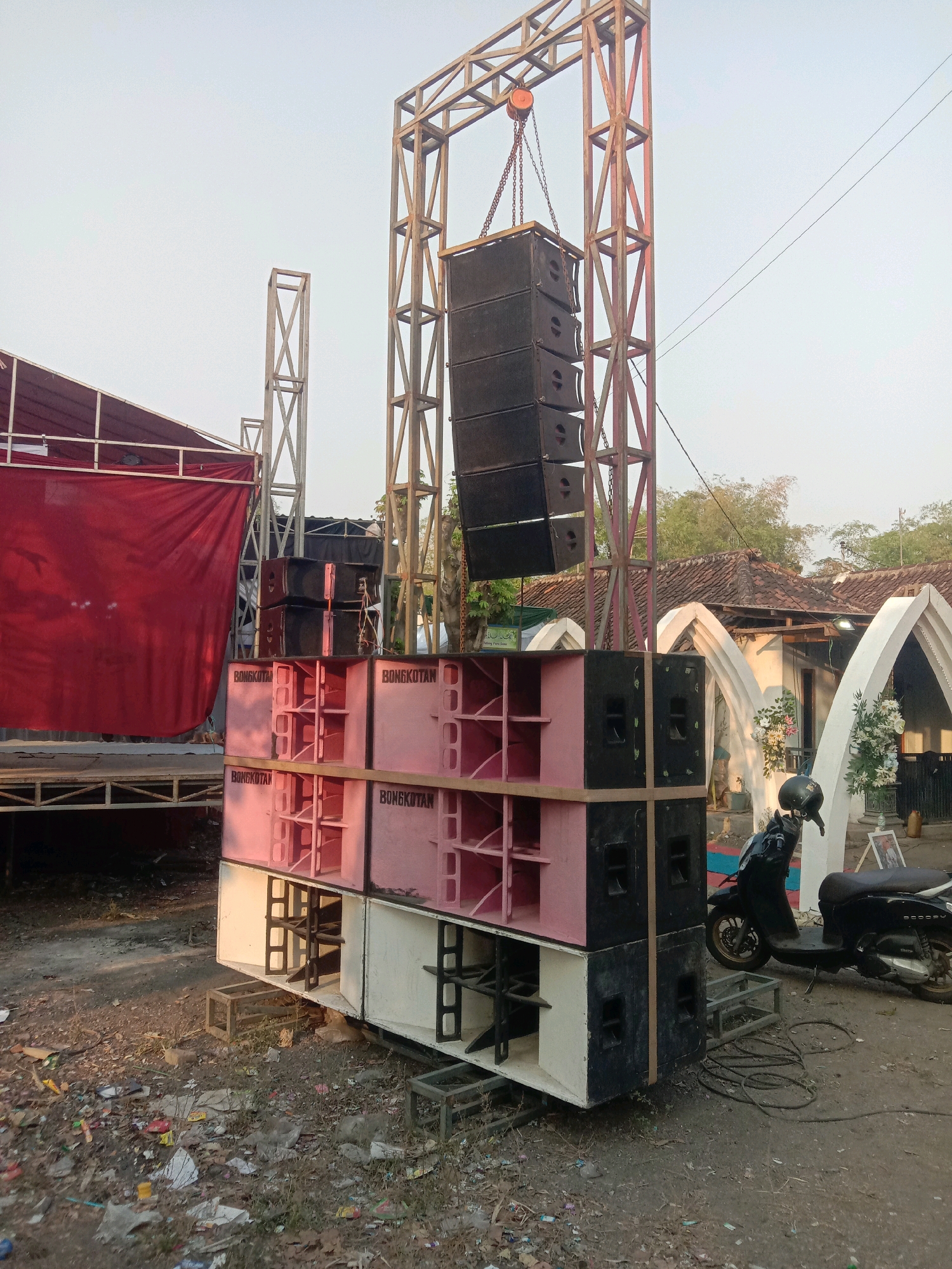 The image depicts an outdoor scene in Karang Anyar, Indonesia, likely before an event. A large sound system is set up, featuring several large speaker cabinets stacked on top of each other. Some of these speakers are pink and labeled "BONGKOTAN," while others are black. A metal truss structure supports additional black speakers suspended from above by chains. To the left, a stage area is visible, draped with a large red cloth. In the background, traditional Indonesian houses with tiled roofs are present. A black scooter is parked to the right of the speaker setup, and various cables and electrical equipment are scattered on the ground. The ground itself is unpaved and littered with debris, including plastic waste and dried vegetation. The overall impression is one of preparation for an outdoor gathering or performance.