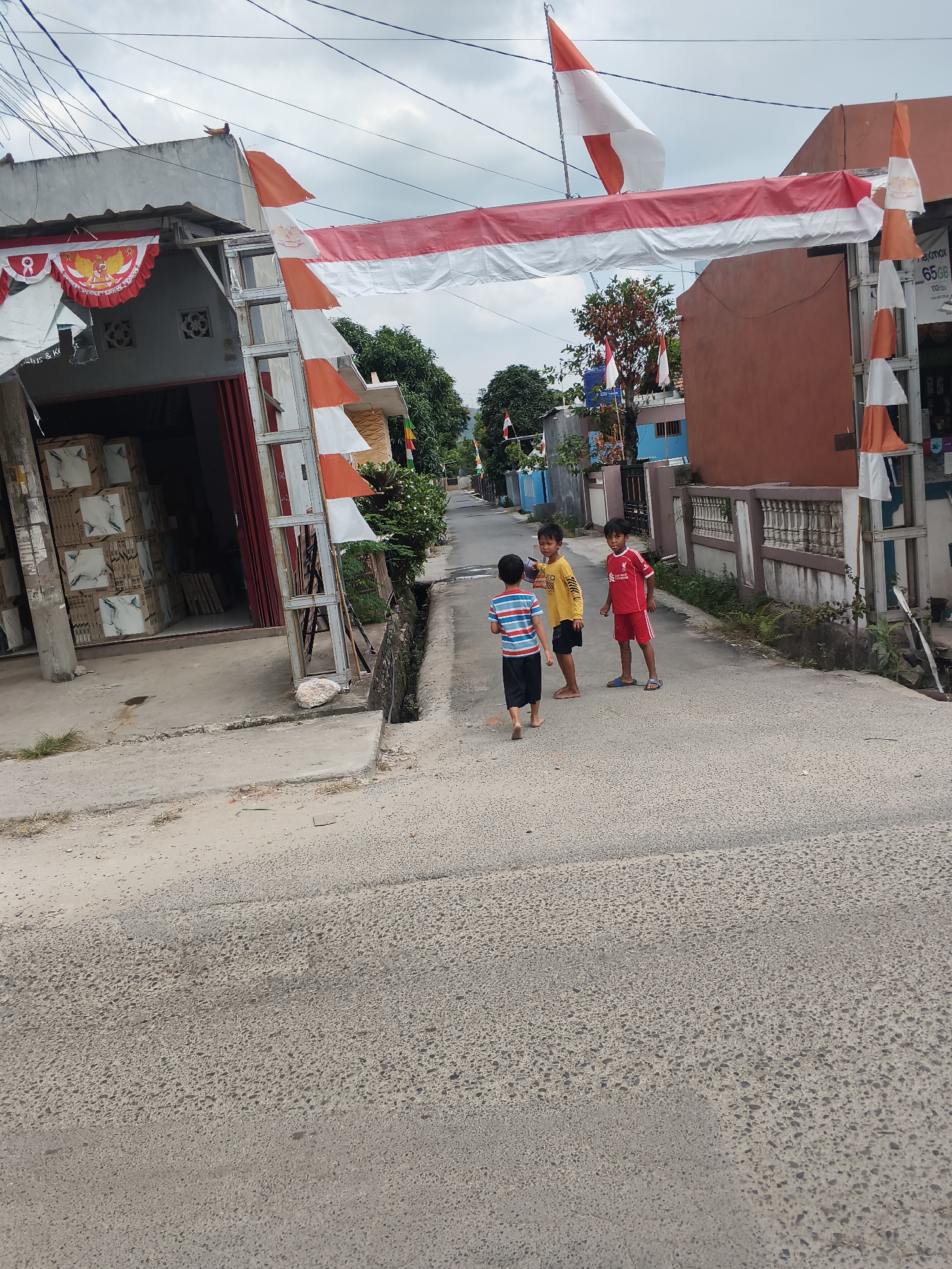 The image depicts a residential street in Bandar Lampung, Indonesia, decorated with Indonesian flags for what appears to be a national celebration. A red and white banner spans across the street, suspended from structures on either side. In the foreground, three young boys are standing on the asphalt road. The boy on the left wears a striped t-shirt and dark shorts, while the middle boy is in a yellow t-shirt and black shorts. The boy on the right is wearing a red Liverpool FC jersey and red shorts. To the left of the street, a building with an open storefront displays stacks of tile boxes. The right side of the street features a sidewalk with a low, decorative wall and some vegetation. Overhead, electrical wires crisscross the sky, which is overcast. The overall scene captures a moment of everyday life in a decorated urban environment.