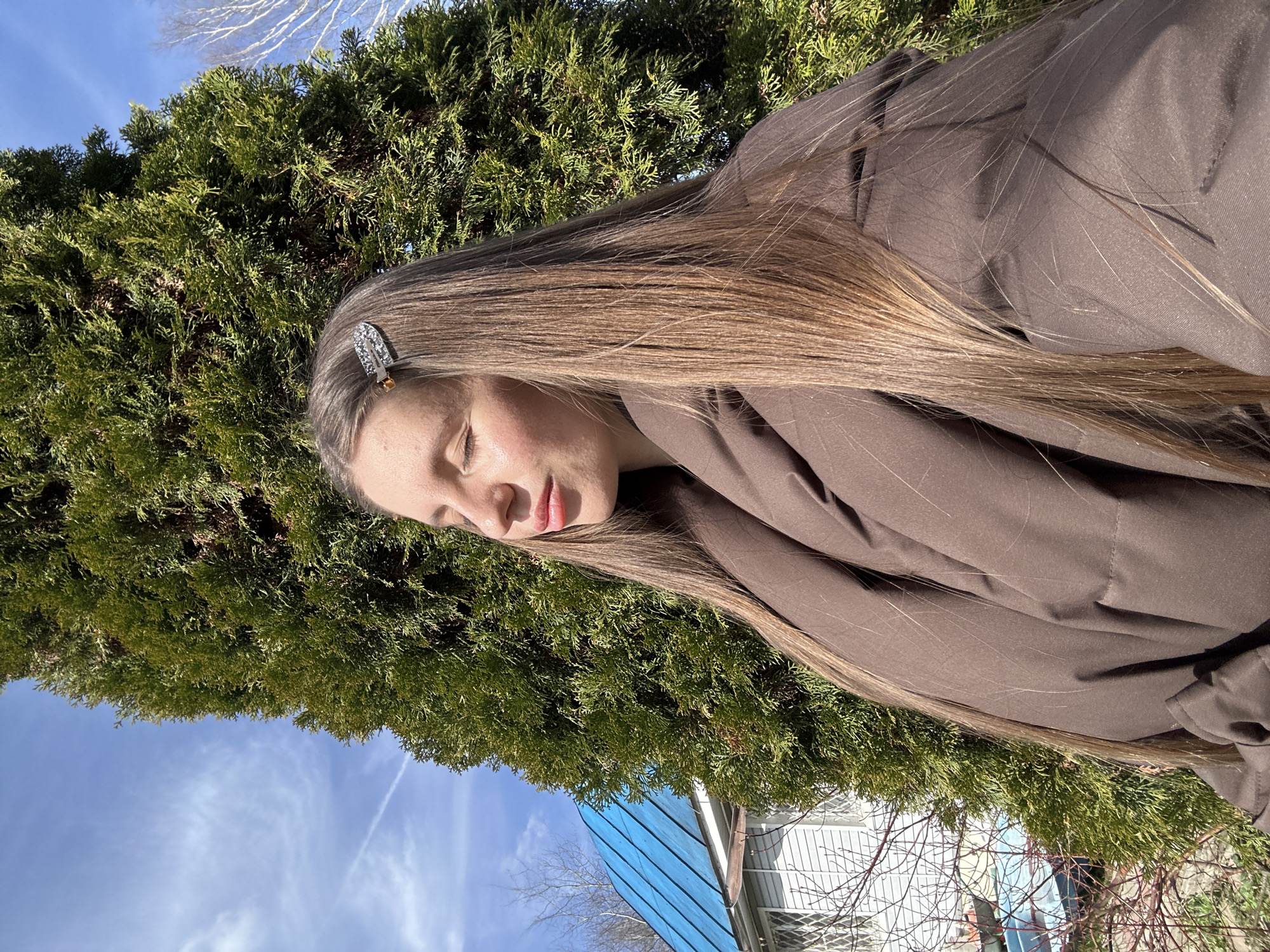 A young woman with long, straight, light brown hair is outdoors. A silver hair clip is visible in her hair. She is wearing a brown jacket or coat. She has her eyes closed. Behind her is a green coniferous tree. A blue sky with some clouds is visible. Parts of a blue roof and bare tree branches are in the background. There is no indication of a location in Мещёры, Russia.