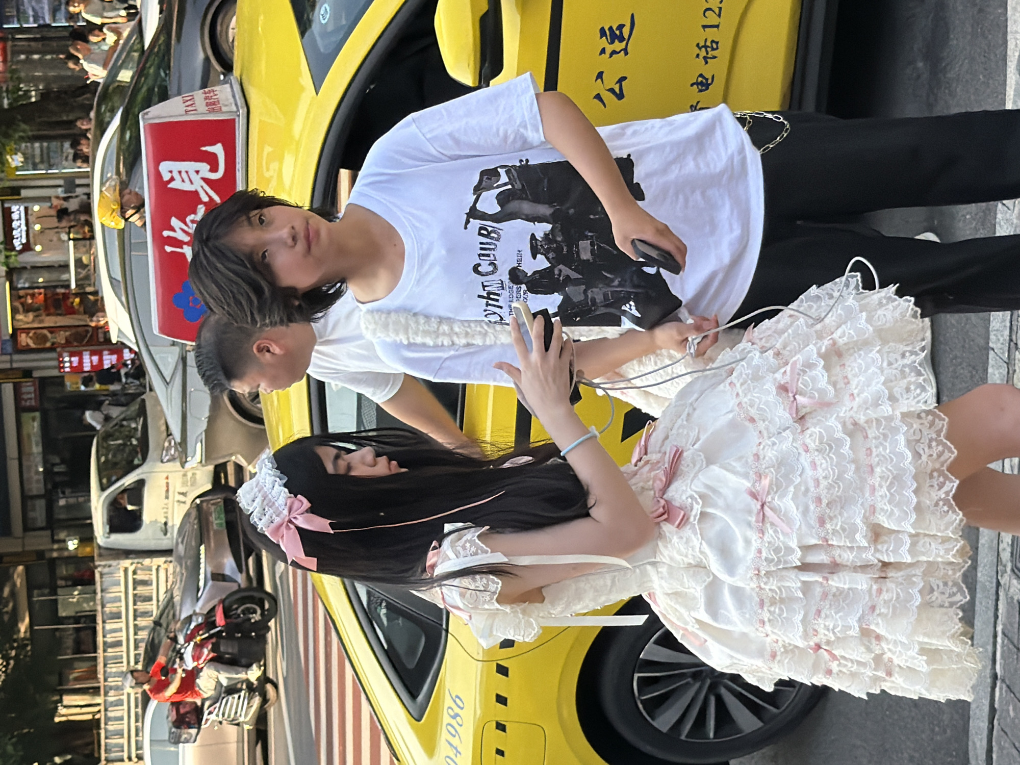 This media file captures a street scene in Yuzhong District, China, with a bright yellow taxi prominently featured. Two individuals are the main subjects. One, a young person with dark hair, is wearing a white t-shirt with a black graphic print and dark pants. They are holding a smartphone. Beside them, a young person with long dark hair is dressed in an elaborate white lolita-style dress adorned with pink ribbons and lace. They are also holding a smartphone.
The scene appears to be outdoors, likely during the day, given the lighting. The background shows a busy street with other vehicles, including a white car and a motorcycle with a rider wearing a red jacket, suggesting urban activity. A sign on the taxi is visible, with Chinese characters and the word "TAXI" in English. Text on the taxi also includes "出租汽车" (rental car) and what appears to be a phone number or code, "电话123". Another piece of text on the taxi's side reads "24986". The overall atmosphere seems casual, with the subjects engaged with their phones, possibly documenting their surroundings or interacting digitally. The Lolita fashion of the second individual adds a distinctive cultural element to the scene.