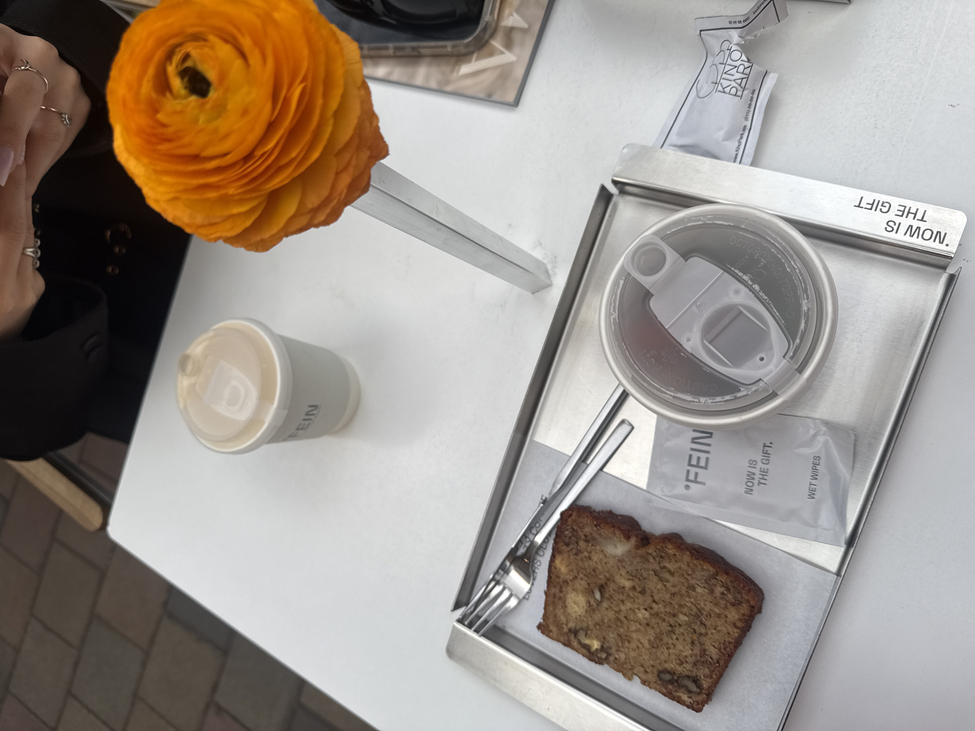 The image depicts a table setting in what appears to be an outdoor café, likely in Yerevan, Armenia, given the presence of "KinoPark.am" printed on a package. On the white tabletop, there is a single orange ranunculus in a simple silver vase, a white disposable coffee cup labeled "FEIN," and a stainless steel tray holding a half-eaten slice of banana bread with a fork, a closed disposable wet wipe package, and a small packet of sugar or sweetener. The tray has the words "NOW IS THE GIFT" etched into its edge. A hand wearing rings is visible in the upper left corner of the image, implying someone is sitting at the table. The coffee cup has a warning in Russian about being hot. The overall environment suggests a contemporary café setting.