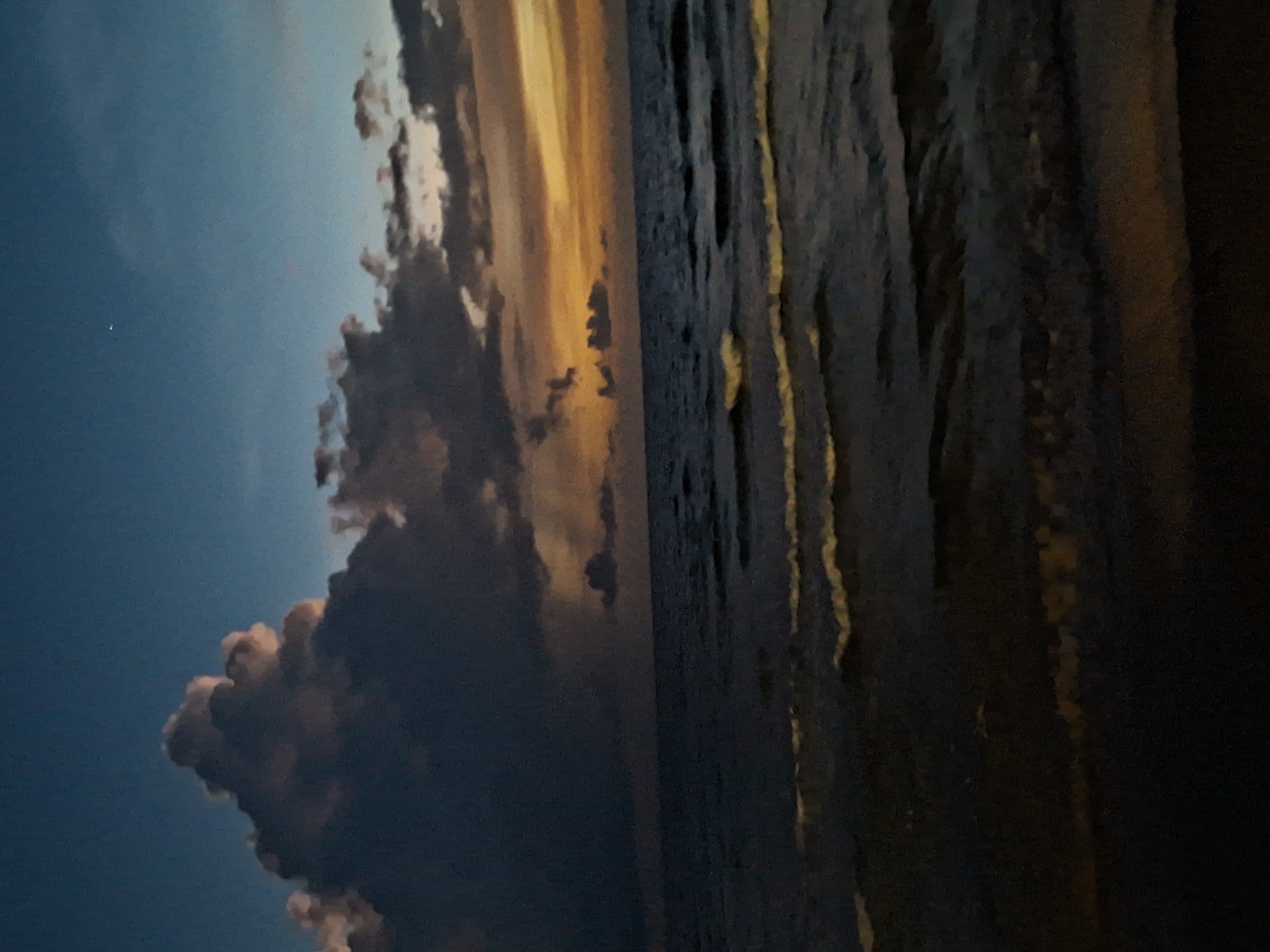 A nighttime photograph captures a serene ocean scene under a twilight sky. The sky transitions from a deep blue at the top to hues of orange and yellow near the horizon, suggesting the last moments of sunset or the early signs of dawn. Dark, dramatic clouds dominate the upper left portion of the frame, with lighter, wispy clouds scattered across the sky. A single, bright star is visible in the upper right quadrant. The ocean is dark and rippling, with gentle waves breaking on an unseen shore. The overall mood is peaceful and tranquil. The image does not contain any people or identifiable structures, and no specific location within a city can be determined.