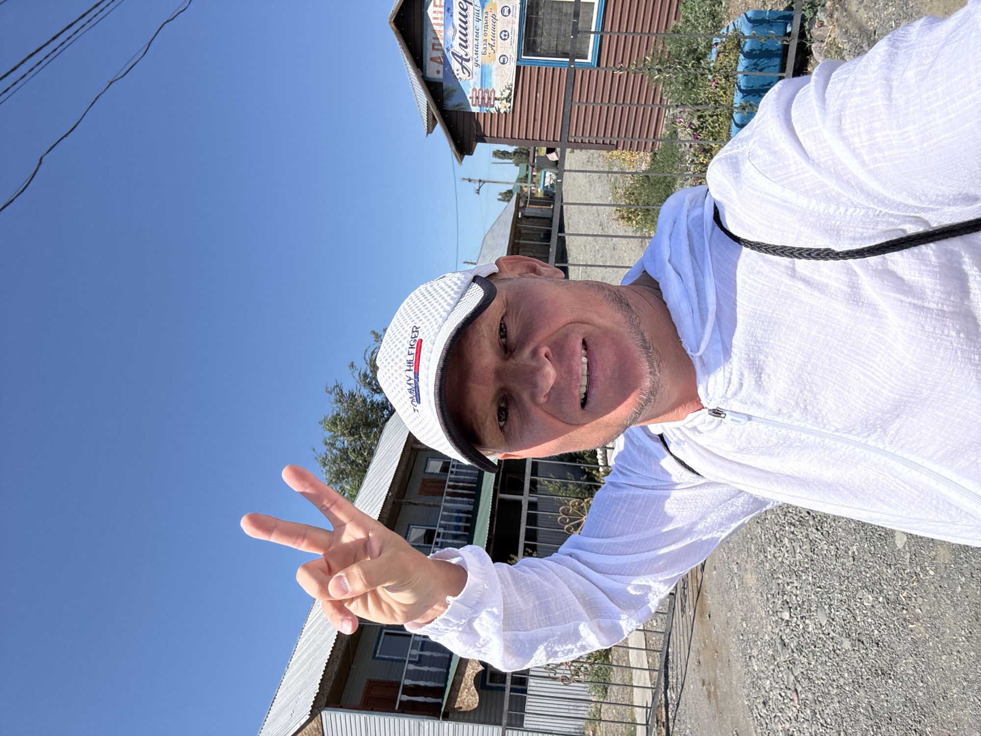 A man wearing a white hoodie and a white baseball cap is taking a selfie outdoors on a sunny day. He is raising his left hand with two fingers extended in a peace sign. In the background, there are several buildings, including a two-story structure with balconies and a smaller, brown-sided building with a sign. The environment appears to be a rural or semi-urban area, possibly a resort or a residential area in Zharbulak, Kazakhstan. The ground is covered with gravel.