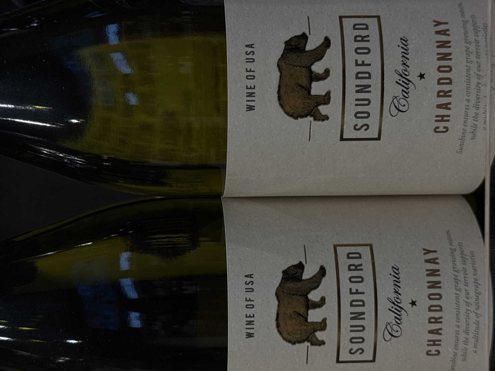 Two bottles of "SOUNDFORD California CHARDONNAY" wine are displayed side-by-side. The labels are light beige with dark brown text and a graphic of a brown bear. The text "WINE OF USA" is prominently displayed above the bear graphic. Below the bear, the brand name "SOUNDFORD" is enclosed in a rectangular box. The region is identified as "California" in elegant script, followed by a star and then "CHARDONNAY" in bold, capitalized letters. A descriptive paragraph about the wine's growing season and terroir is printed below the varietal name, though it is partially obscured and difficult to read in full. The bottles are made of green glass and appear to be in a retail or storage setting. There is no discernible connection to Moscow, Russia, as the context is a product label that specifies "WINE OF USA" and "California."