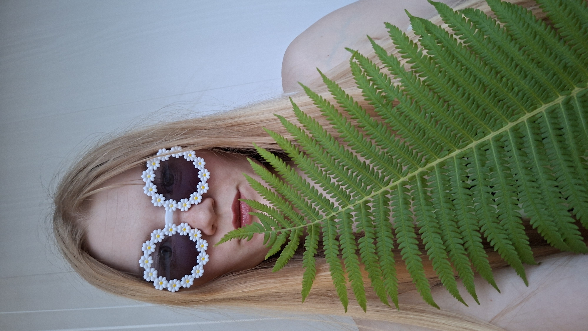 A fair-skinned person with long blonde hair is positioned in the center of the frame, holding a large, green fern frond in front of their face. The person is wearing white, oversized sunglasses adorned with small, white daisy-like flowers. Their lips are slightly parted and colored with a light pink lipstick. The background is a plain, light-colored wall, suggesting an indoor setting. The lighting is even, casting no significant shadows. The image is a medium shot, focusing on the upper body of the individual and the fern. There is no contextual information to suggest a specific location within the city of Chaikovsky, Russia.